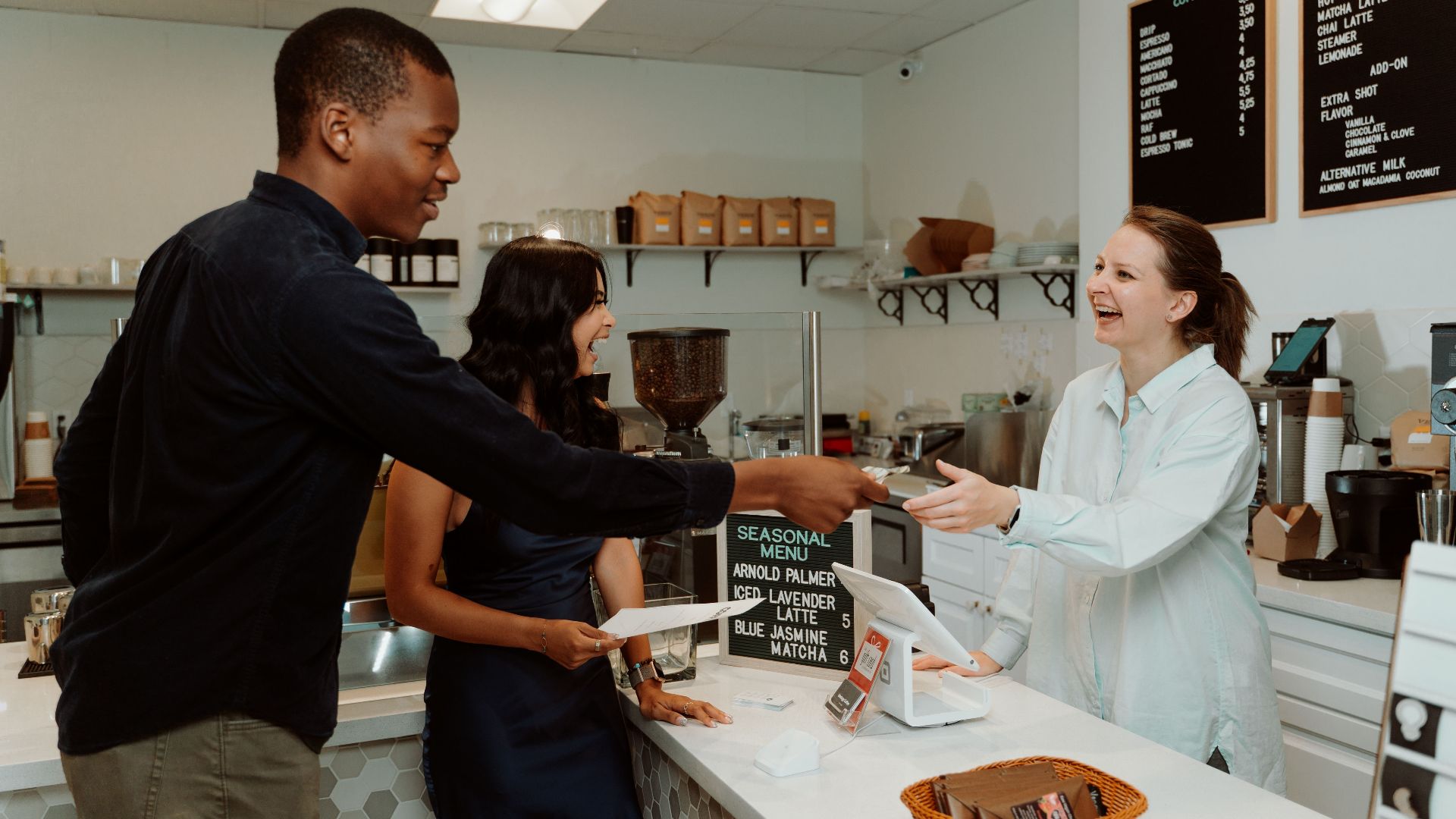 Customers and barista laughing and exchanging payment at a coffee shop counter.