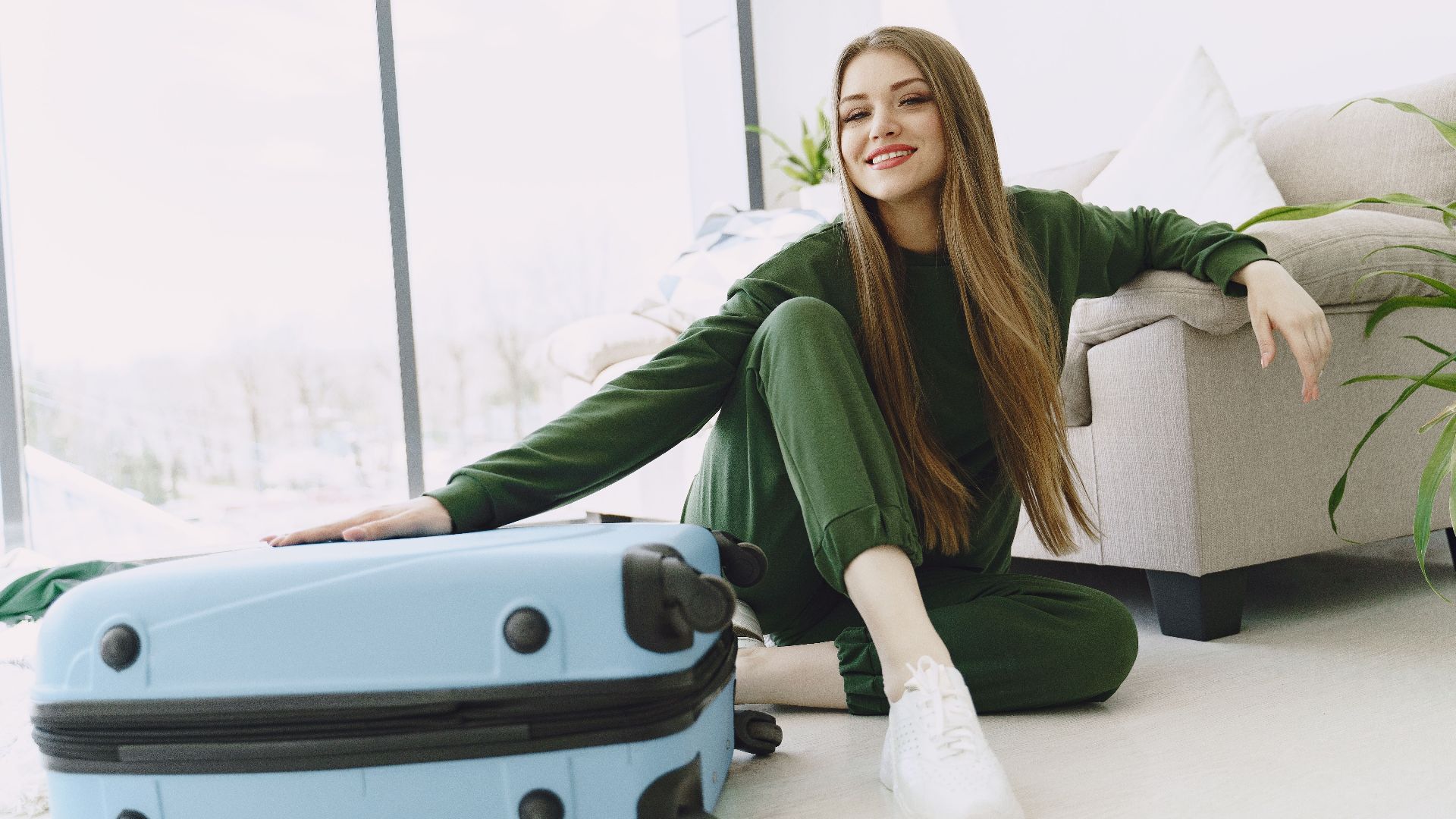 Cheerful female traveler in green sport suit sitting on floor with crossed legs leaning on cozy sofa while putting arm on blue suitcase in contemporary flat with glass wall and looking at camera with smile
