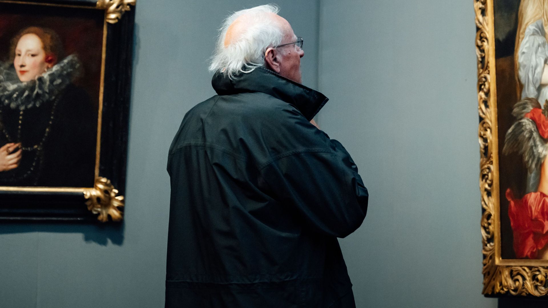 A senior man observes classical paintings in an art museum gallery.