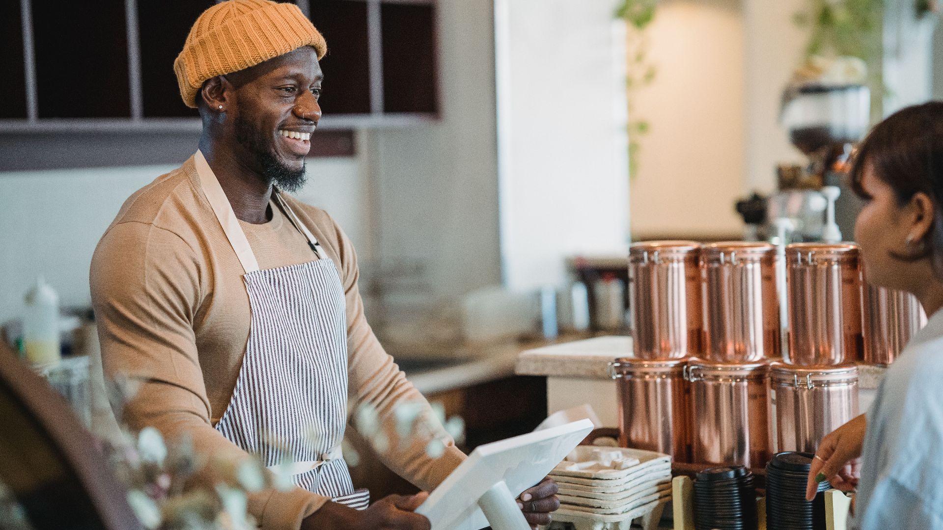 Smiling barista wearing an apron and beanie engaging with a customer at a café counter.