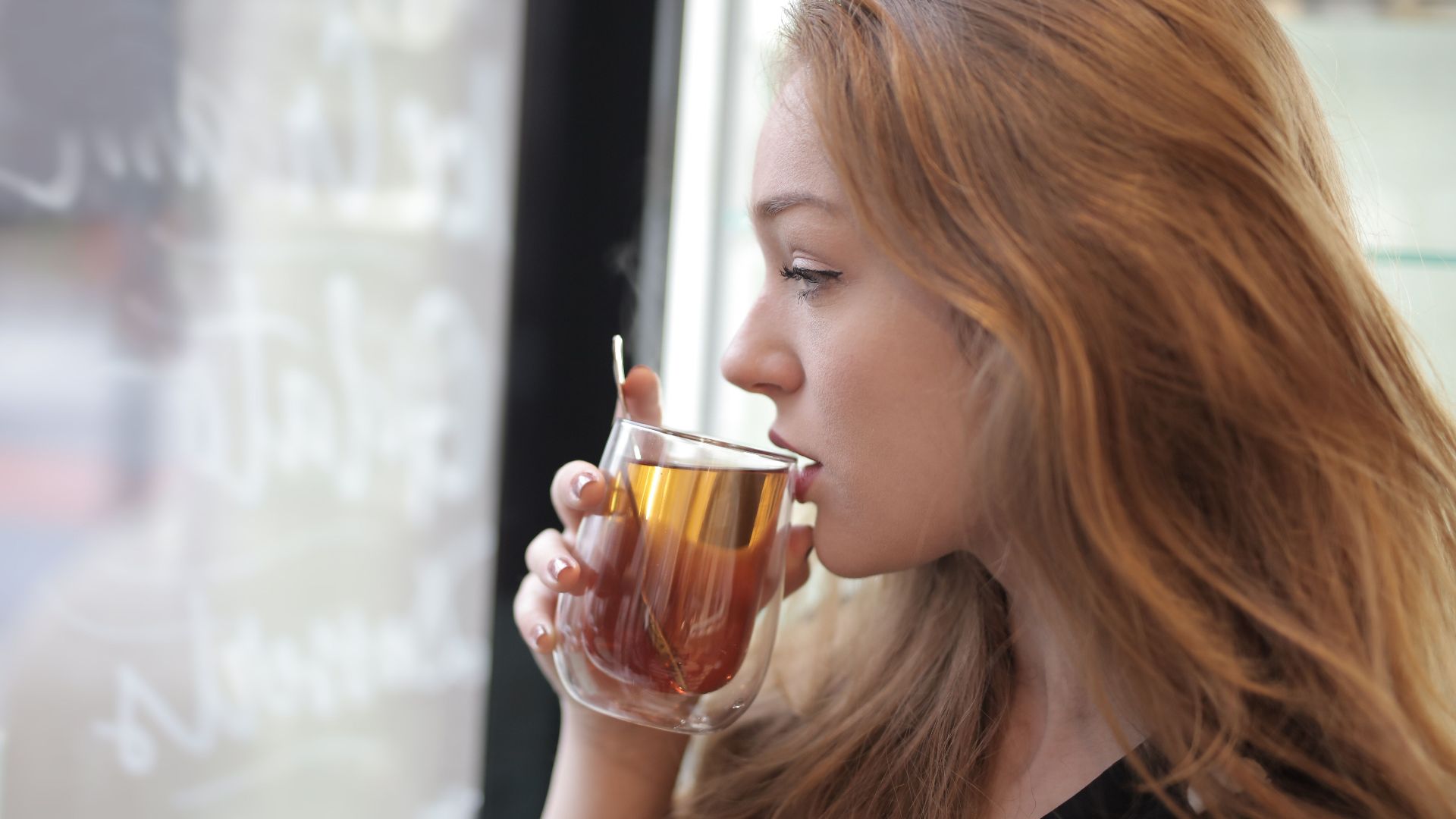 Side view of thoughtful female sipping tasty hot beverage from glass while standing in cafeteria and looking out of window