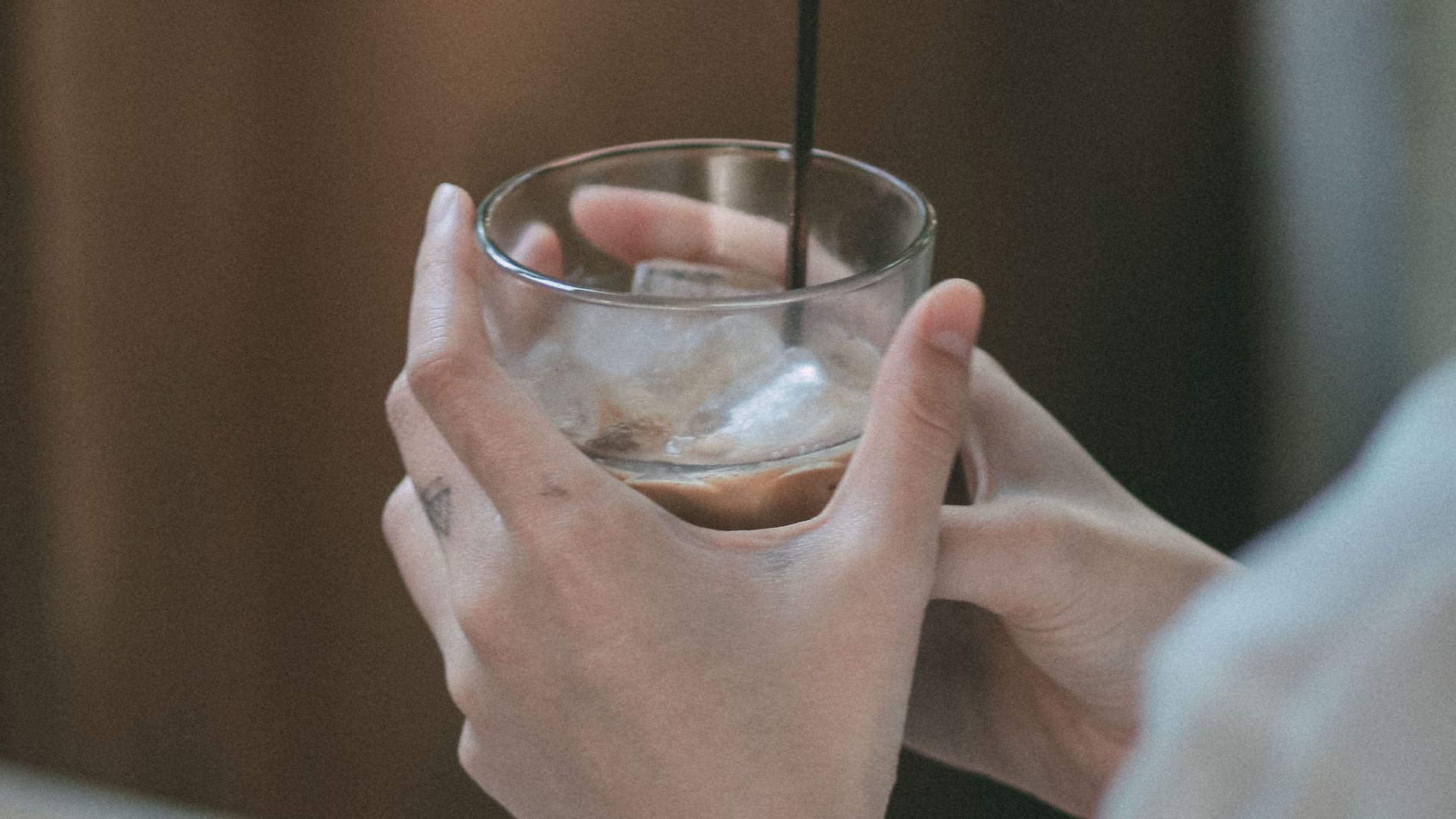 A close-up of hands holding an iced drink in a glass with a blurred background, evoking a relaxed mood.