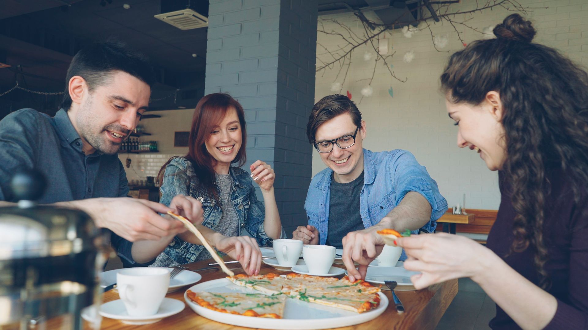 Group of friends cheerfully enjoying pizza and coffee in a cozy café setting.