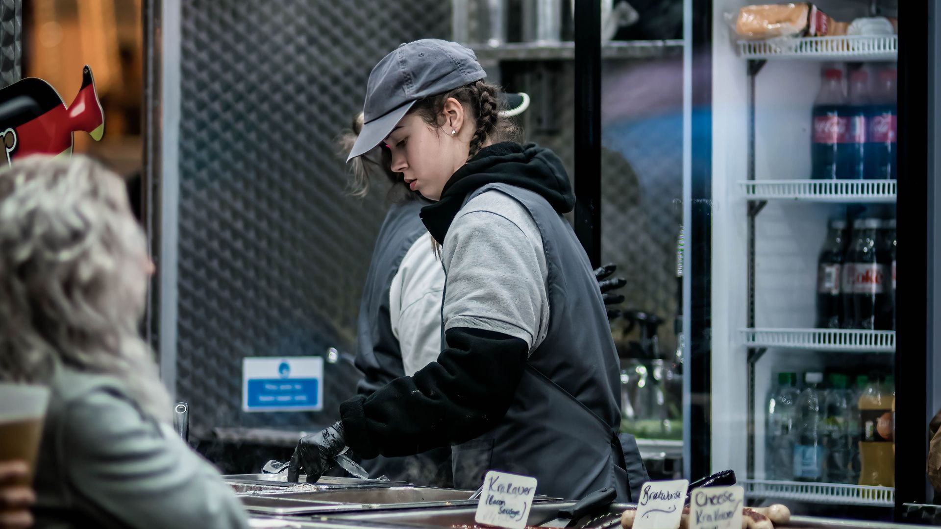 Vendor serving food at an outdoor market stall with cold drinks displayed.