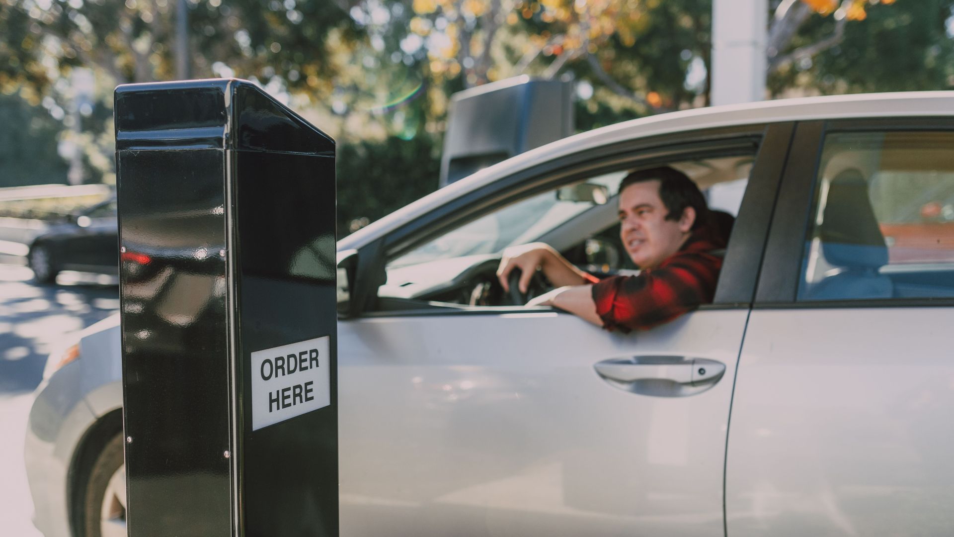 A man in a car orders at a drive-thru window, surrounded by outdoor scenery.