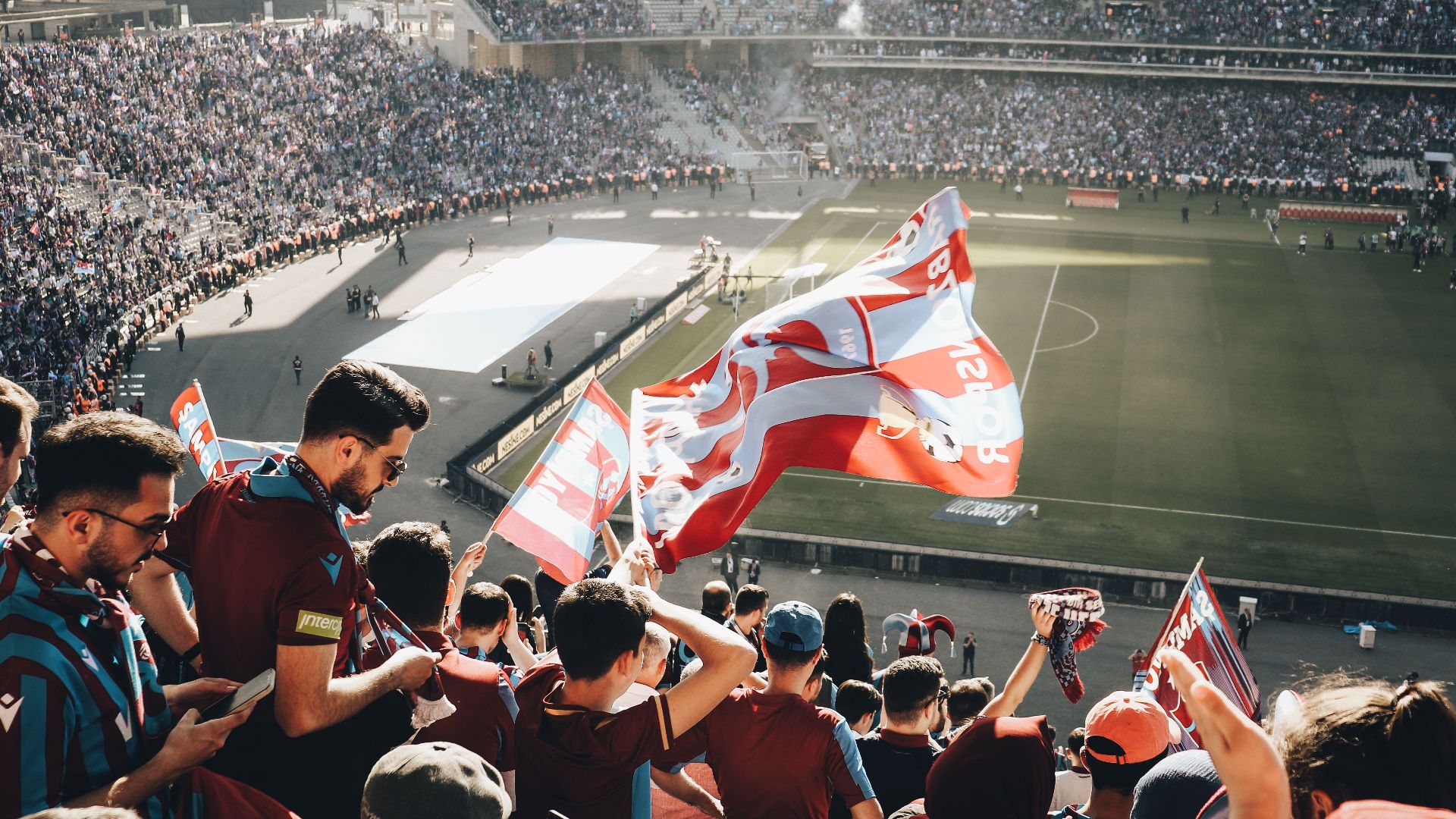 Vibrant crowd with flags cheering at a football stadium during the day.