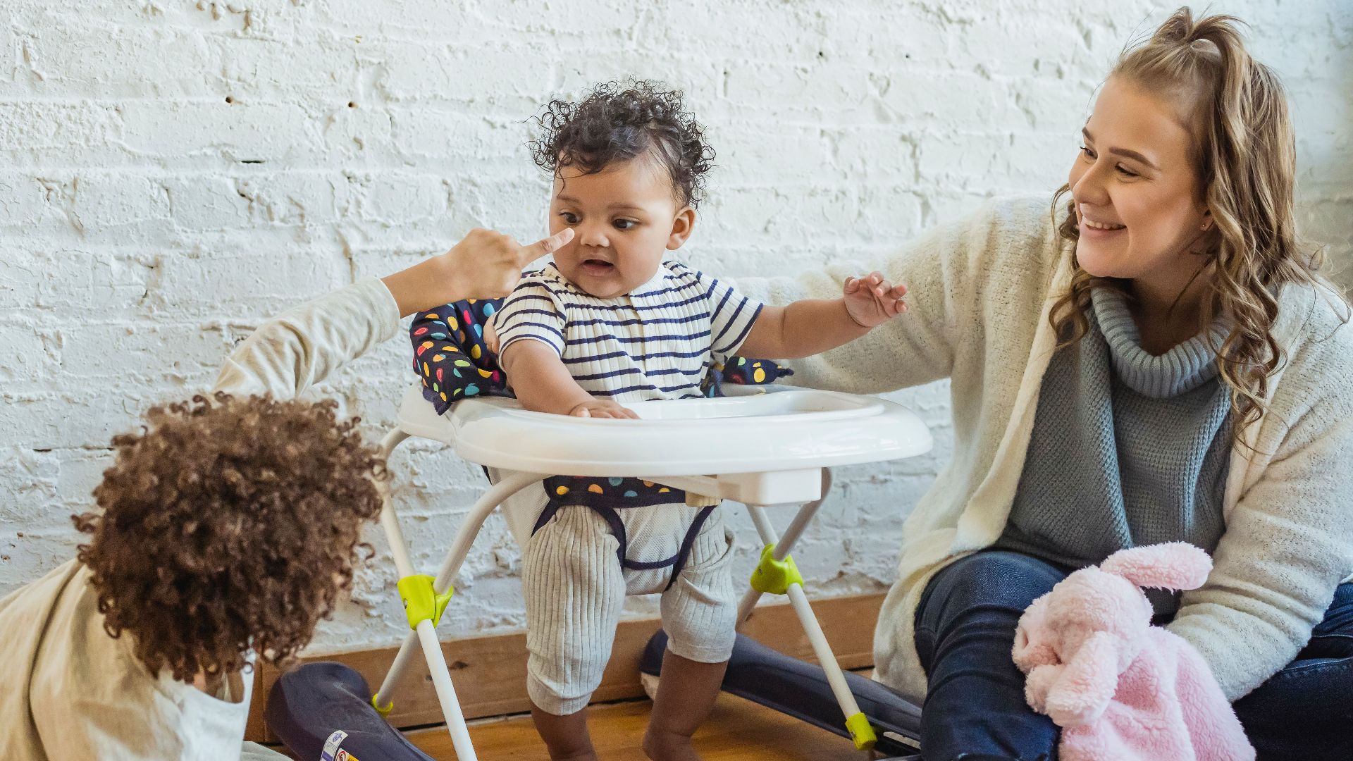 Happy mother with toy playing with black toddler in baby walker and boy while sitting on floor near wall at home
