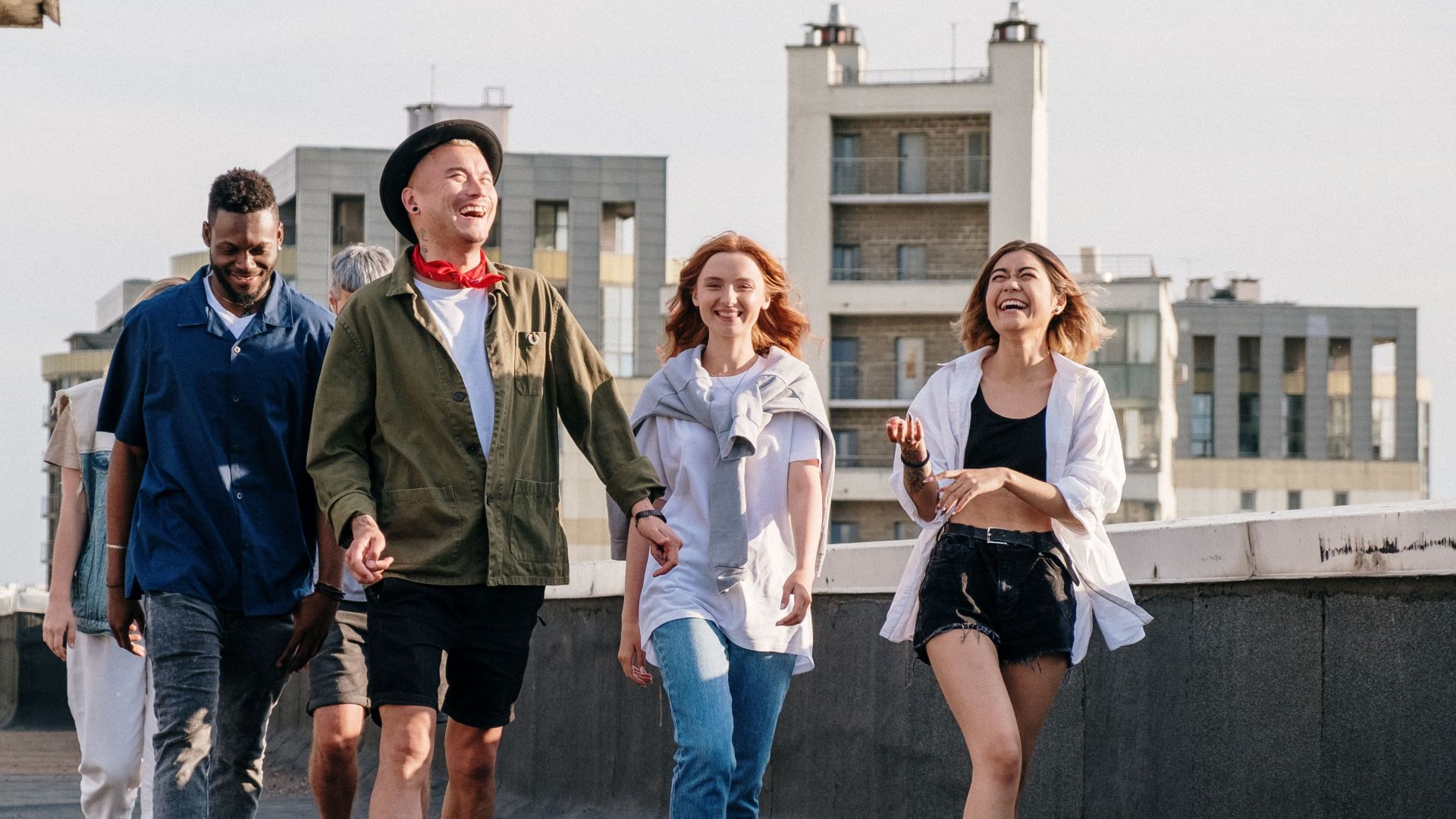 Diverse group of friends laughing and walking on an urban rooftop, enjoying a sunny day.