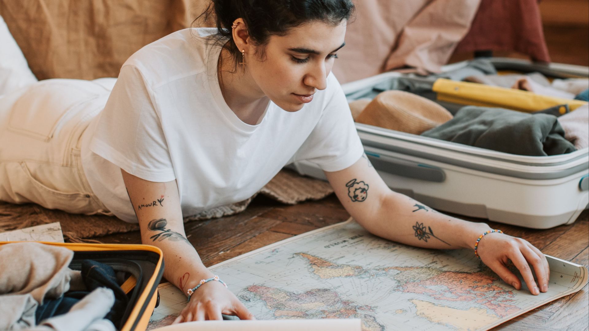 Tattooed woman in white shirt studies map near suitcase on wooden floor indoors. Travel preparation ambiance.