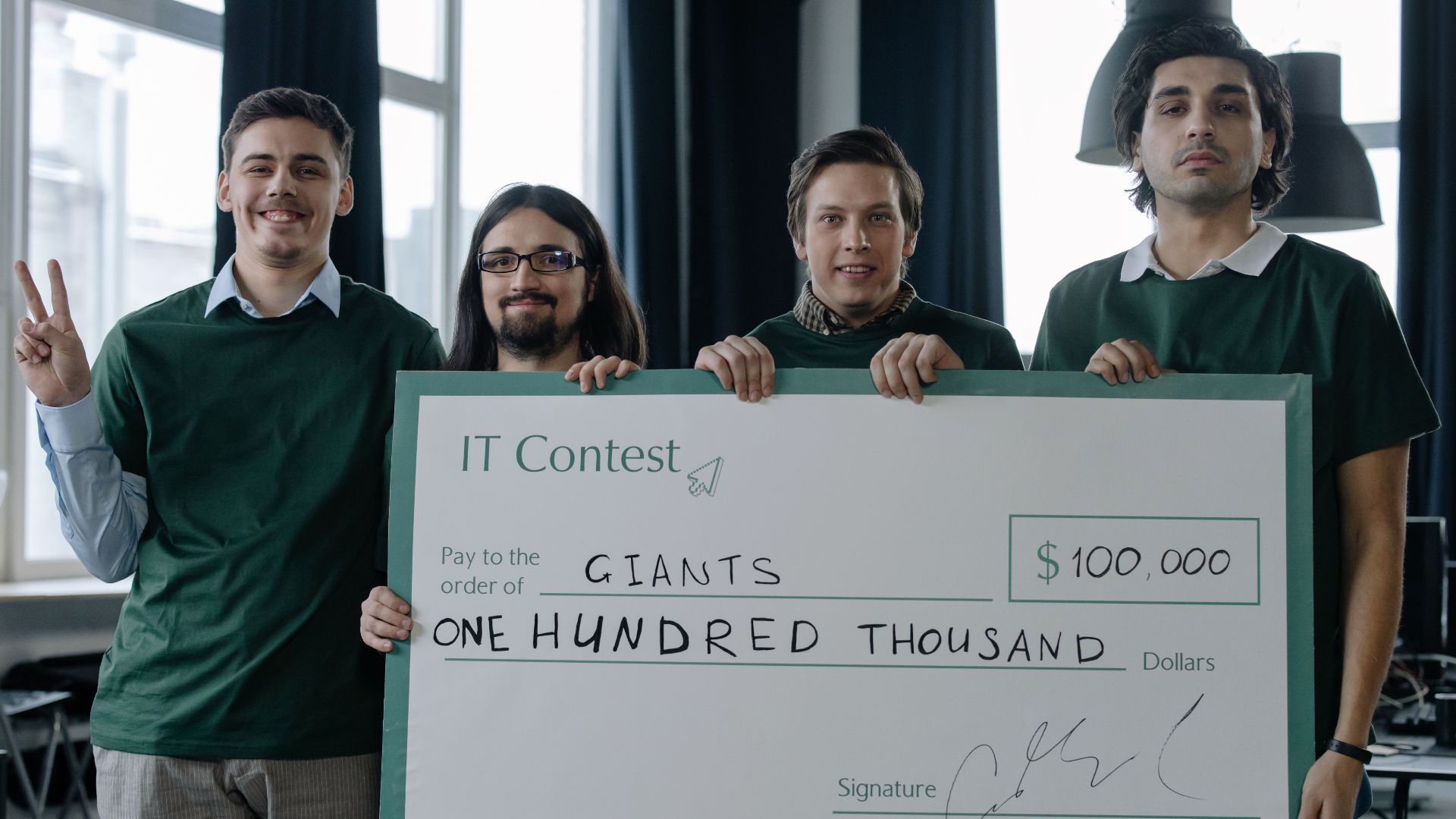 Four men celebrate a $100,000 IT contest victory holding a large cheque inside a modern office.
