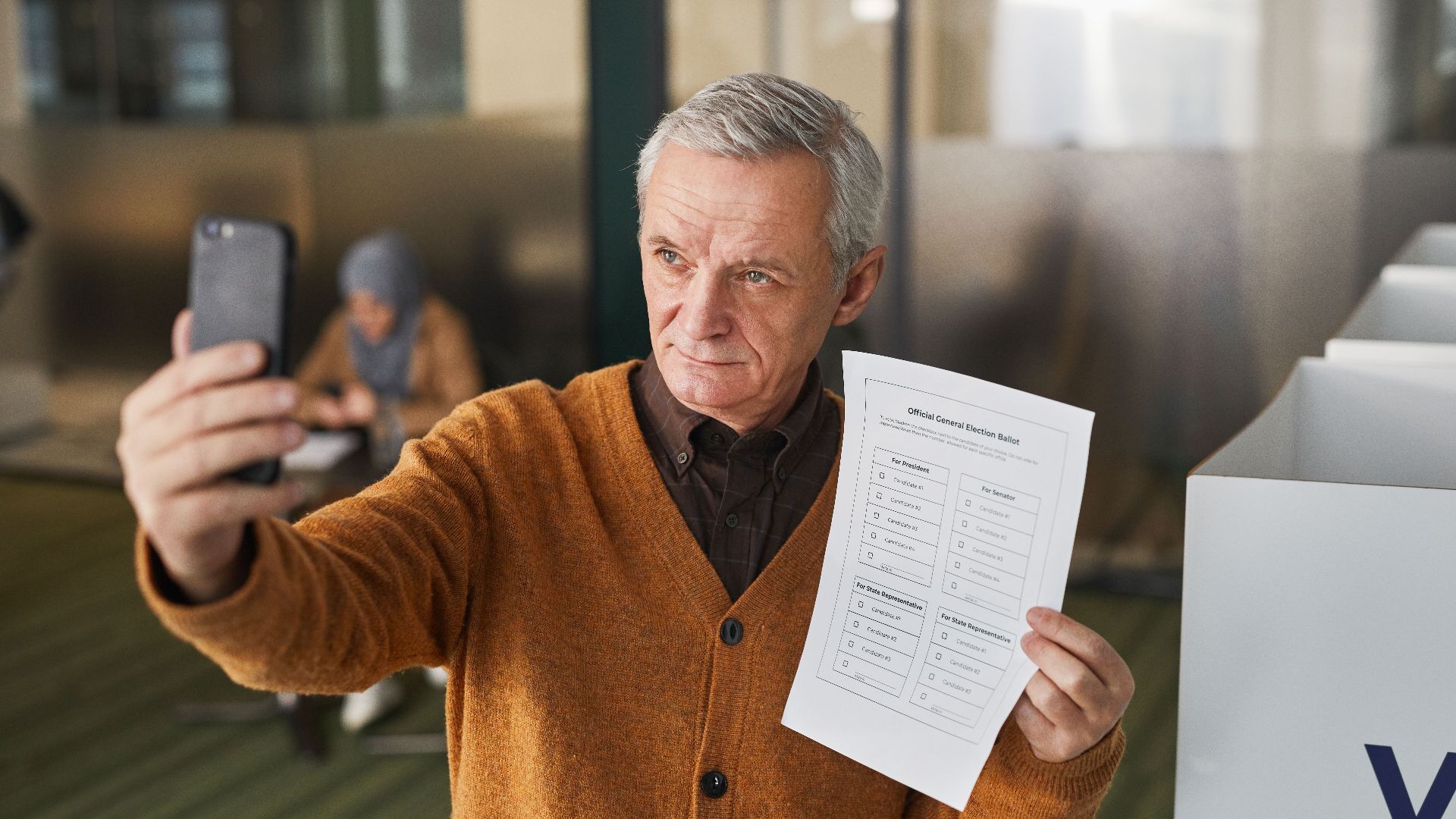 Elderly man with gray hair holding a ballot and taking a selfie in a voting booth.