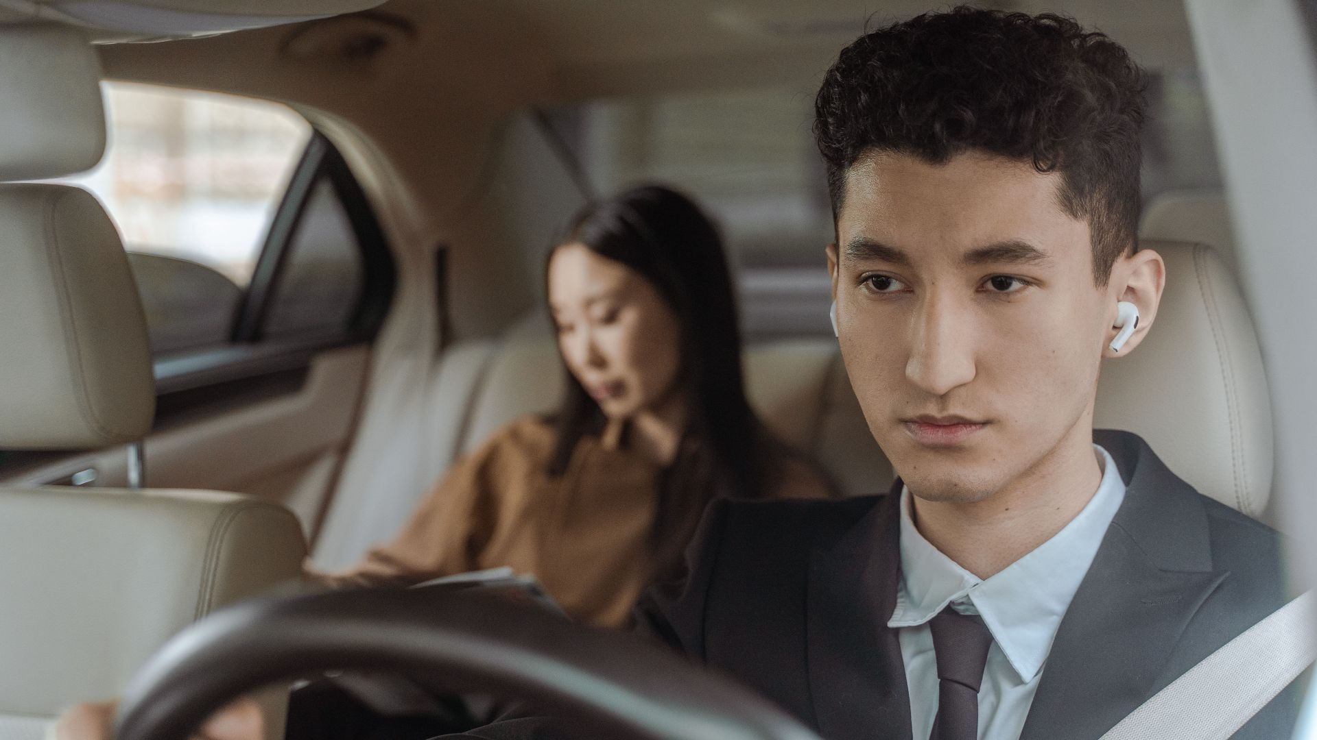 Young professional driver wearing suit and earpiece in luxury car interior with female passenger.