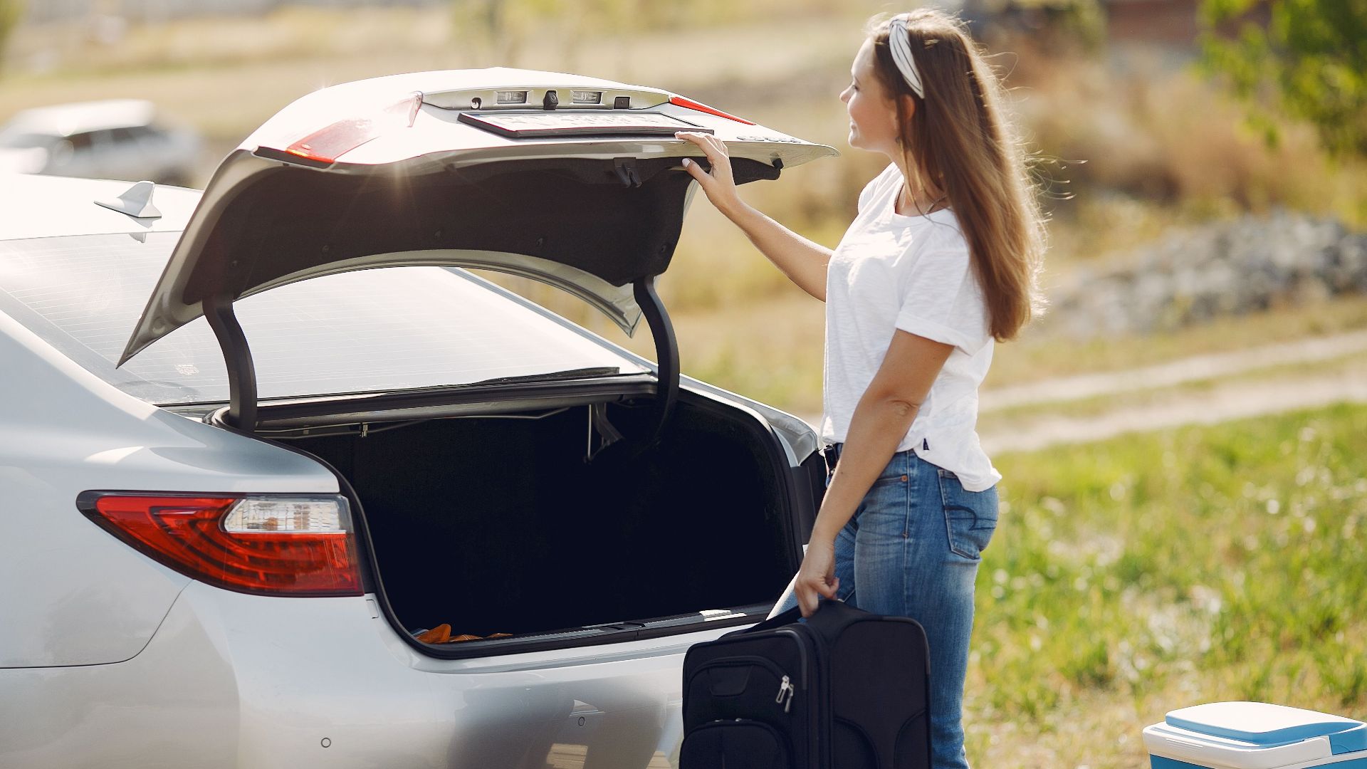 Side view of cheerful female traveler in headband and casual clothes putting luggage in open trunk of modern car while spending summer weekend in countryside