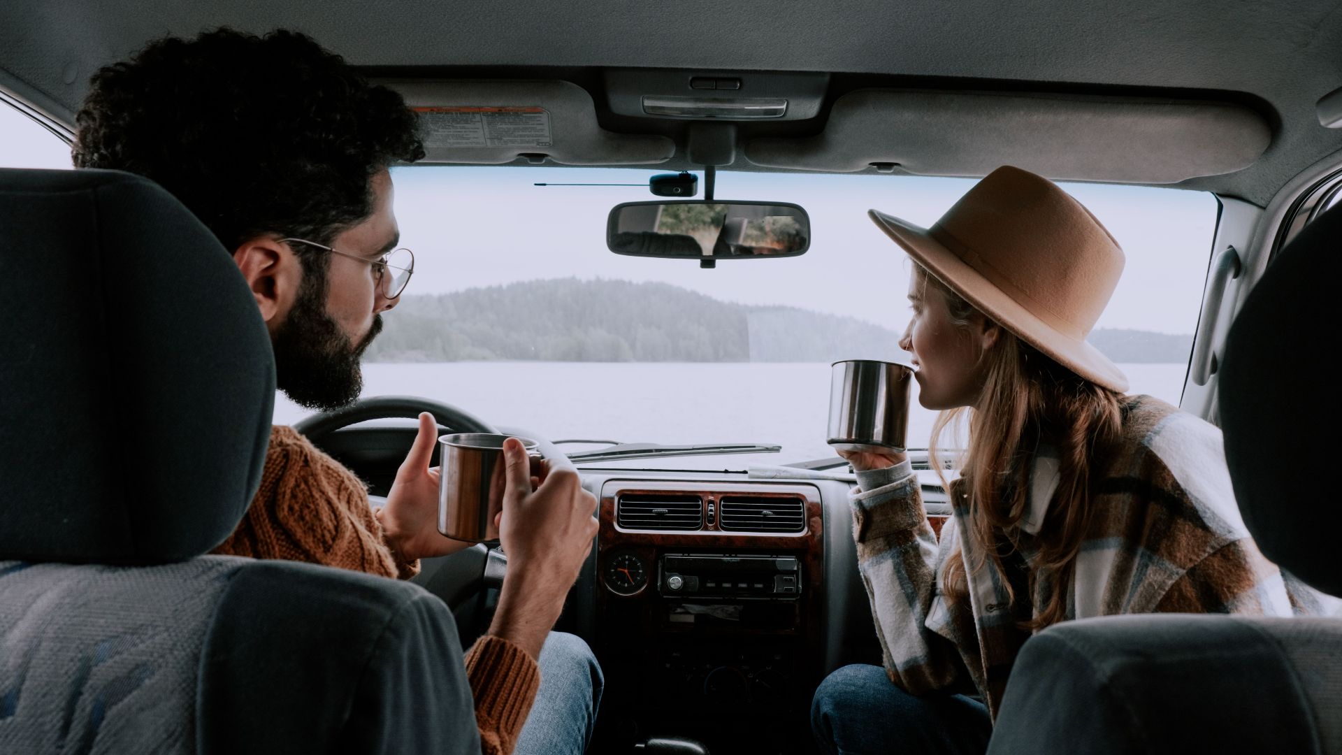 Couple enjoying hot drinks inside car with scenic lake view. Perfect for travel and lifestyle themes.