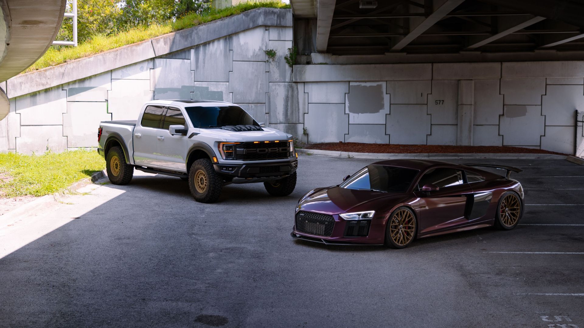 Silver truck and red sports car parked under a highway overpass in an urban setting.