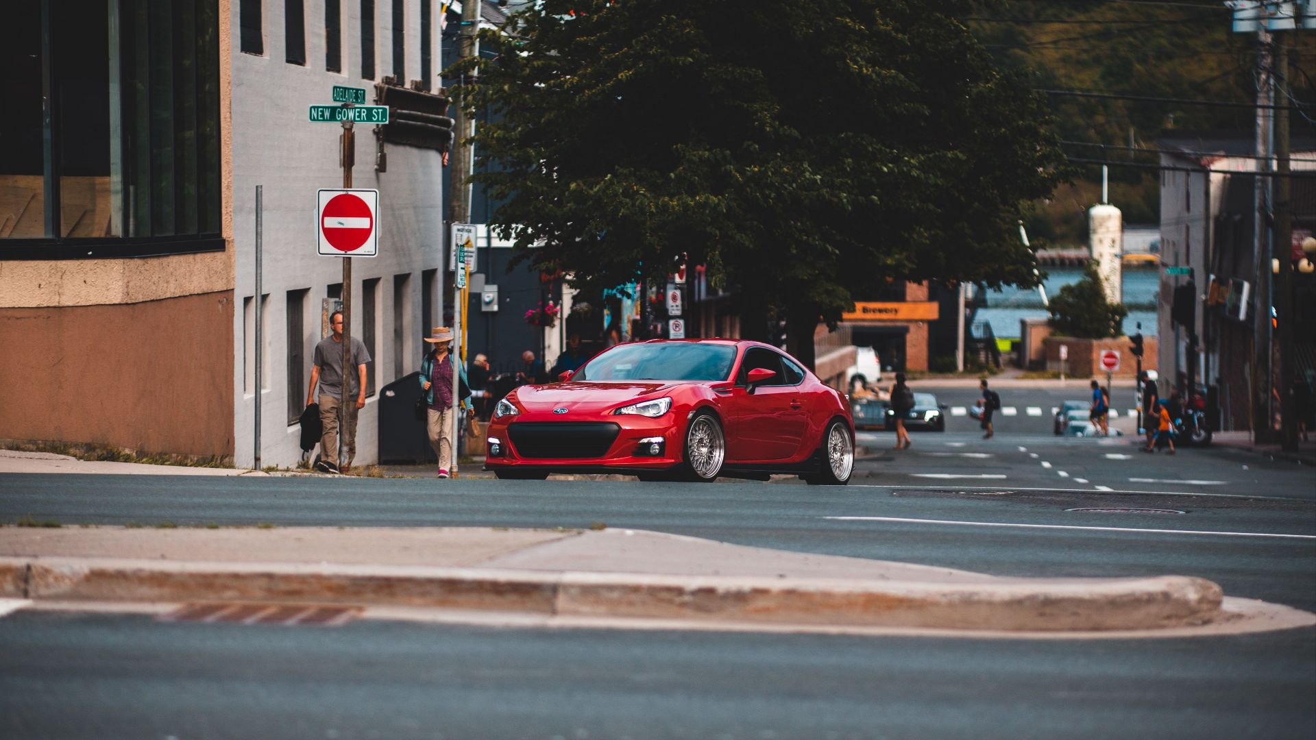 Red sports car turns corner on city street with pedestrians nearby. Urban scene with city signs.