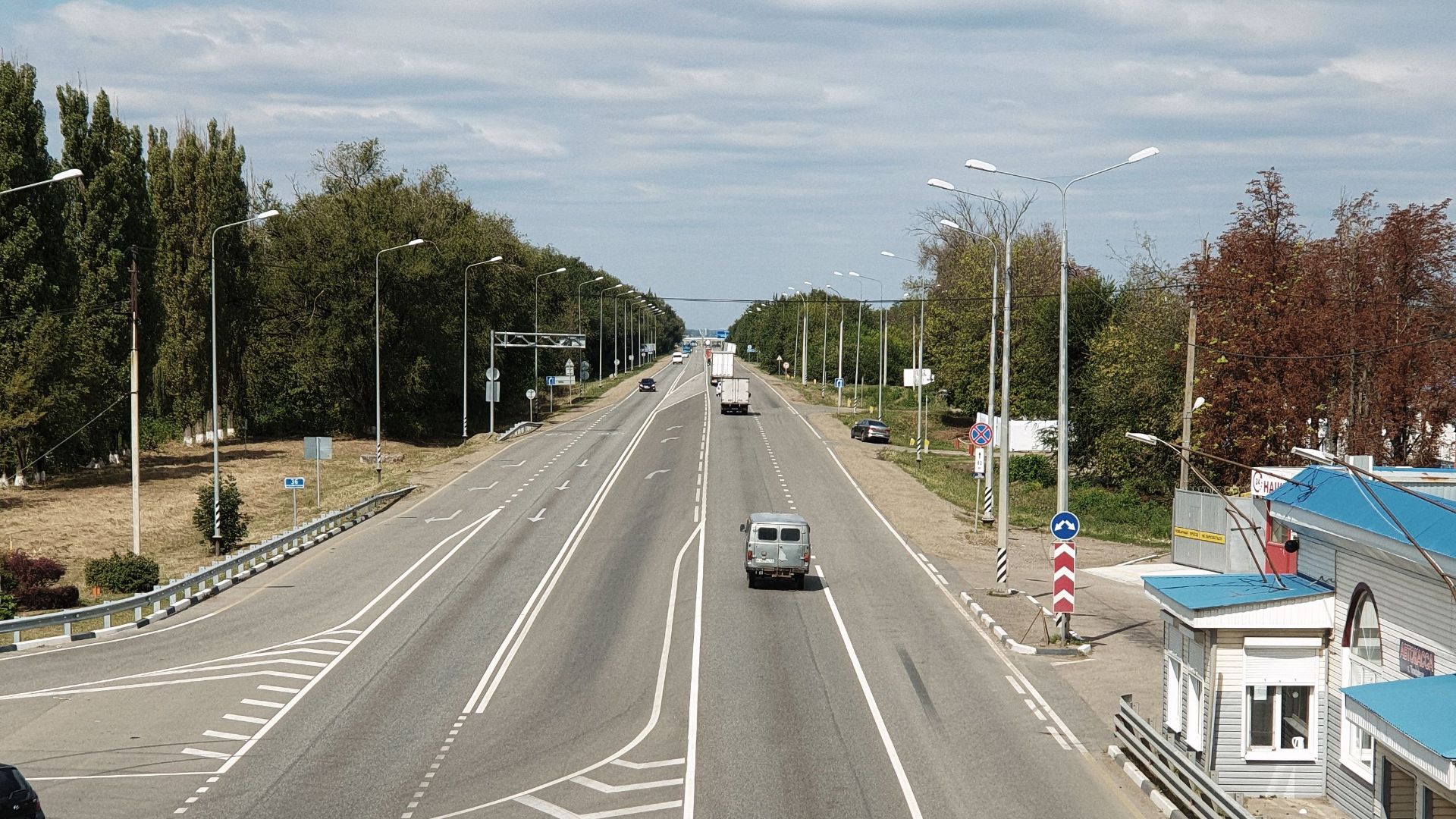 A drone captures an aerial shot of a wide, empty rural road with sparse vehicles surrounded by trees.