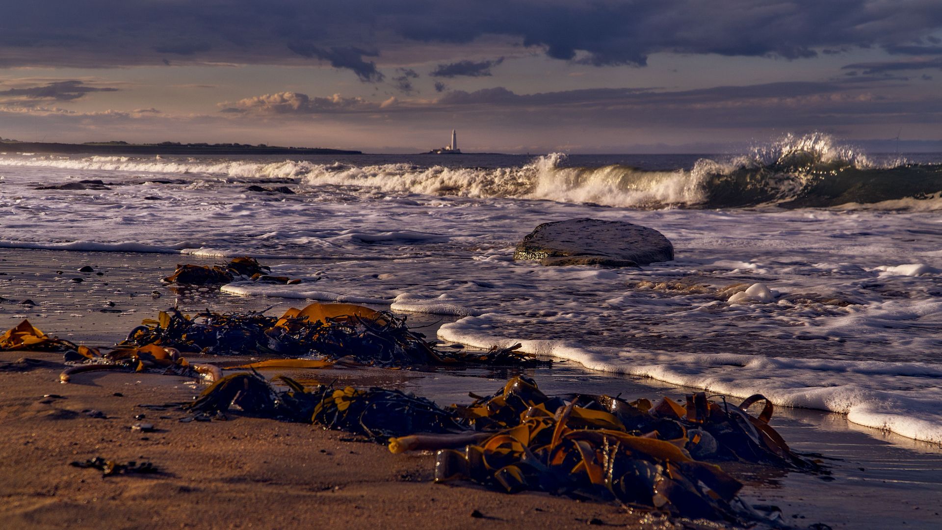 Vibrant waves crash against the shore at sunset, framed by distant lighthouse views.