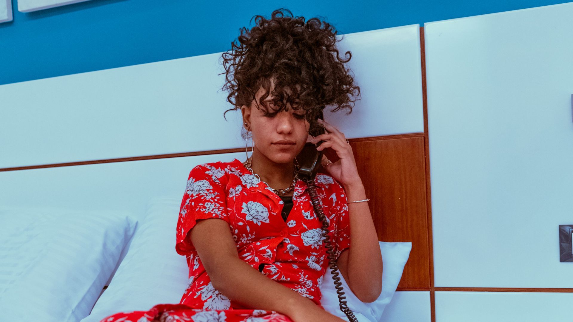Young woman in vibrant red dress talks on hotel phone while sitting on bed.