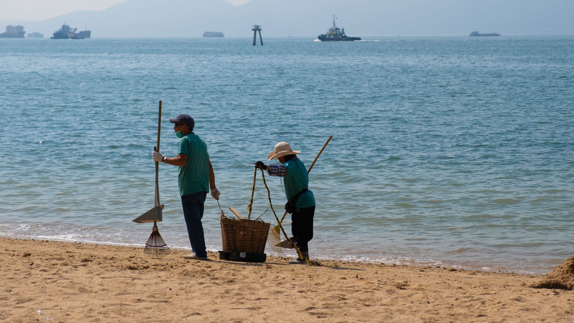 Two workers cleaning a sandy beach with brooms and dustpan beside a calm ocean.