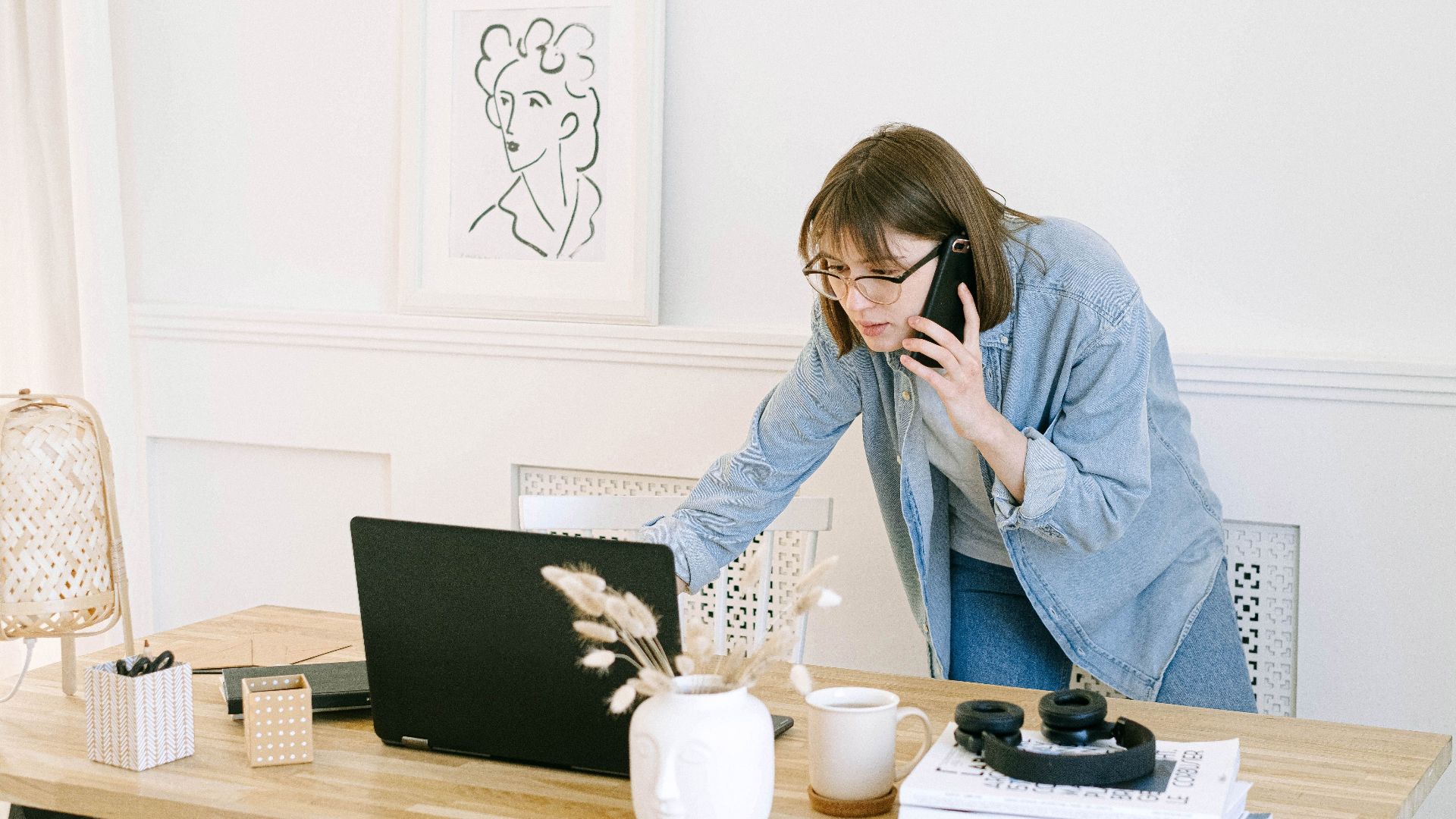 Woman multitasking with laptop and phone in modern home office setting.