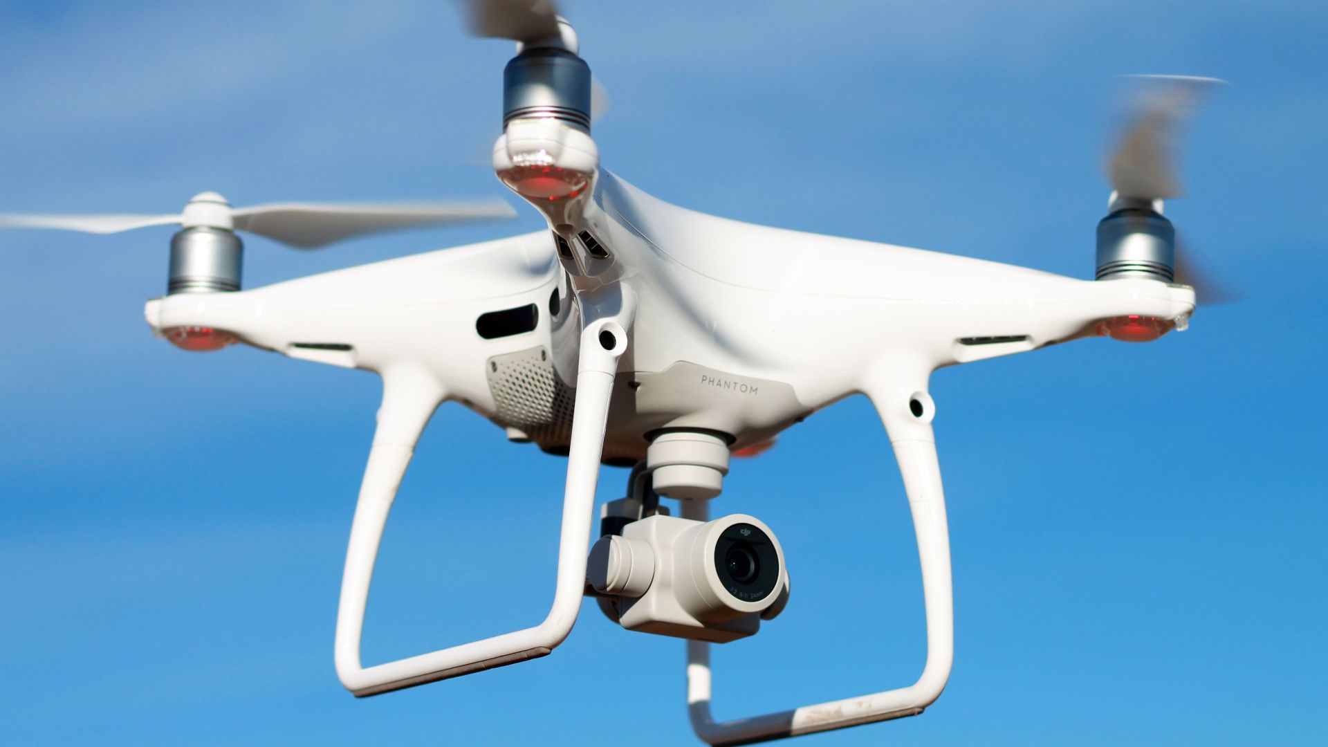 A white drone flying against a clear blue sky, capturing aerial views with its camera.