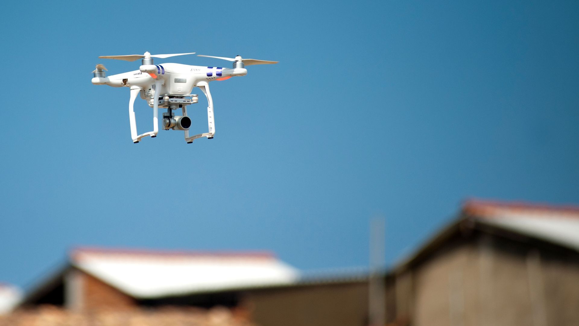 Aerial drone with camera hovering over rooftops beneath a clear blue sky, showcasing modern technology in action.