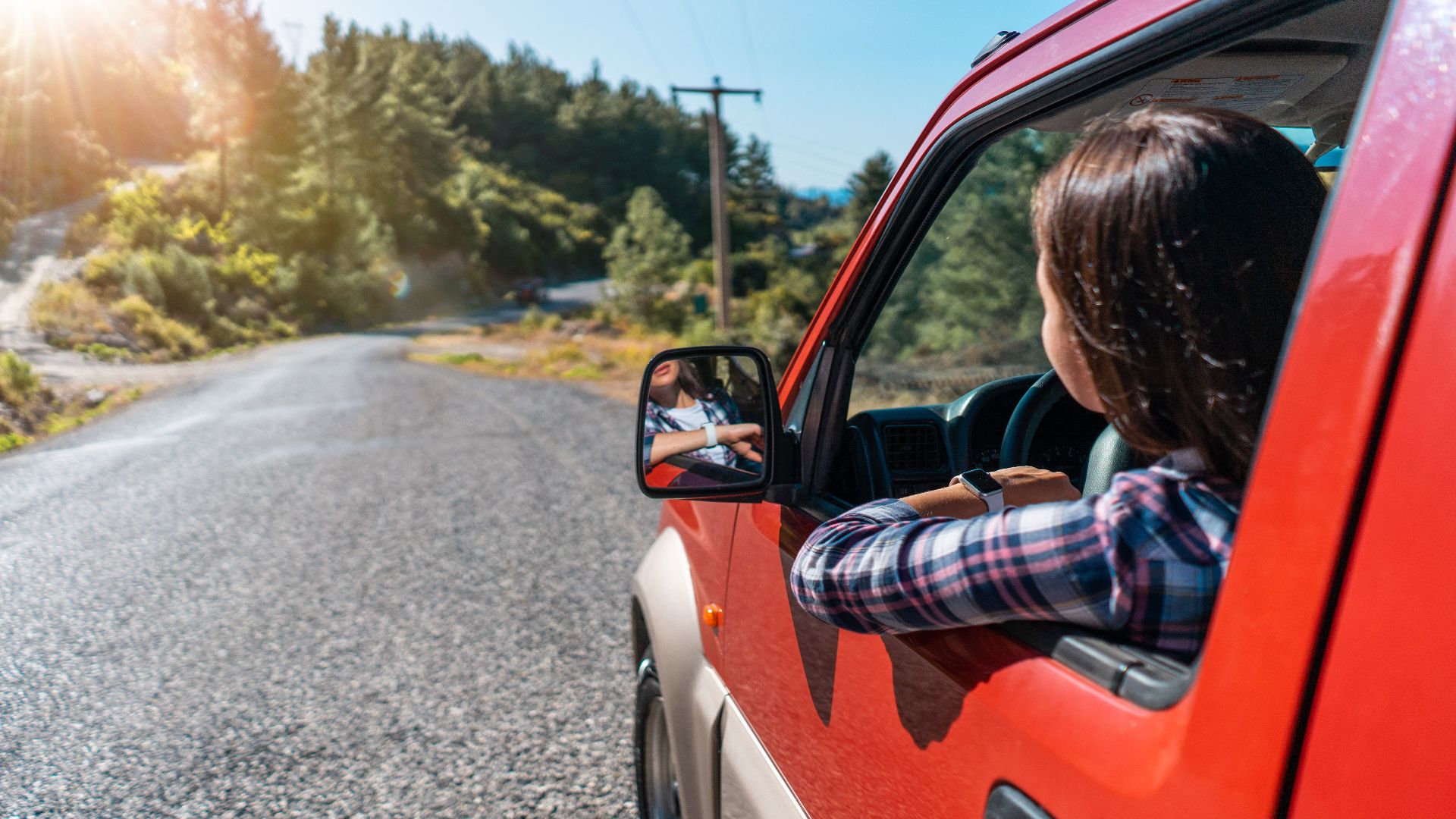 Back view of unrecognizable female driver looking out of window while riding red automobile on asphalt road near green trees