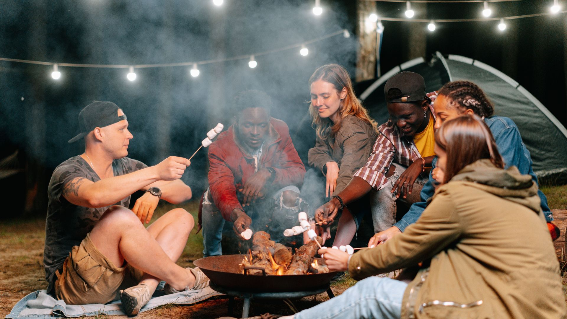 A group of friends relaxes by a bonfire, roasting marshmallows under string lights on a camping trip.