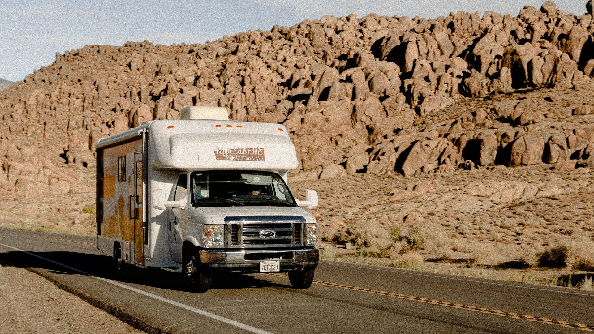 RV driving through a scenic desert highway with rocky mountains under a clear sky.