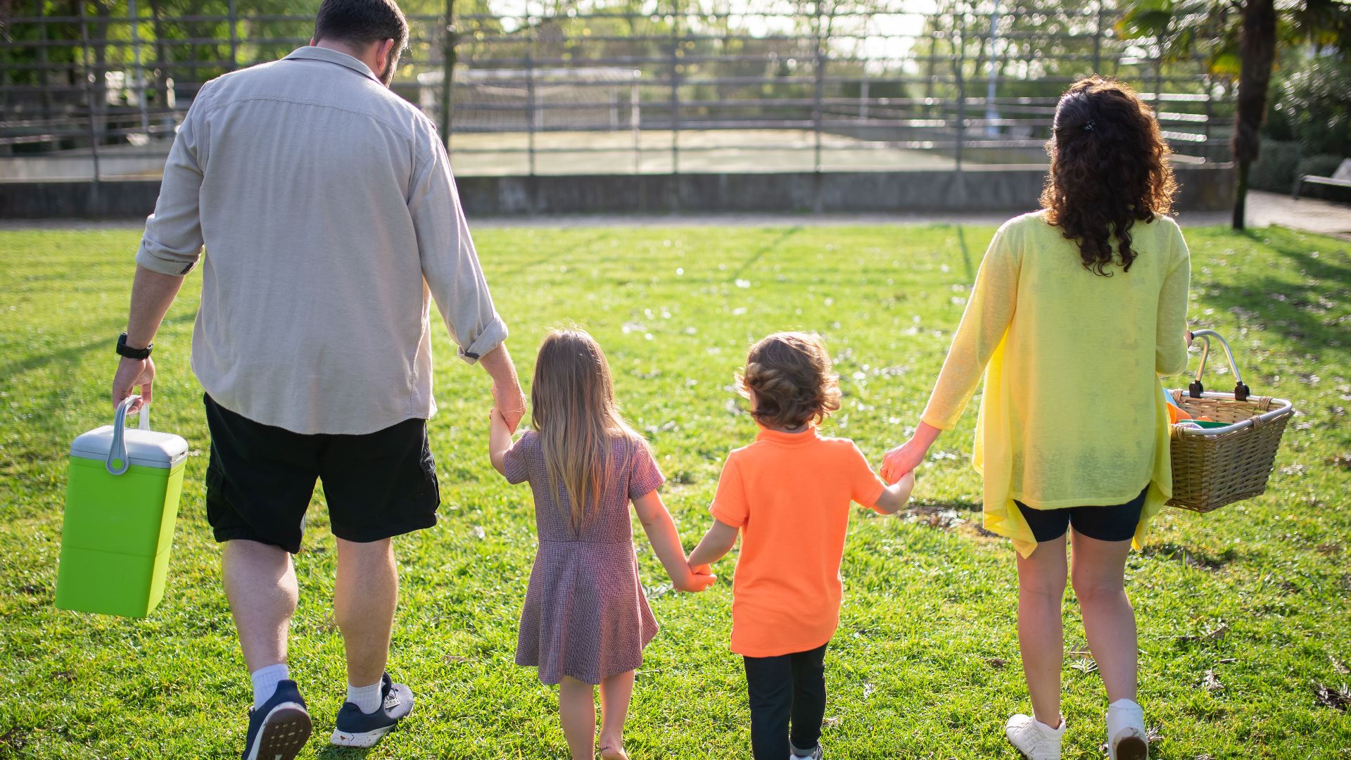 A family of four walking hand in hand through a sunny park carrying picnic supplies.