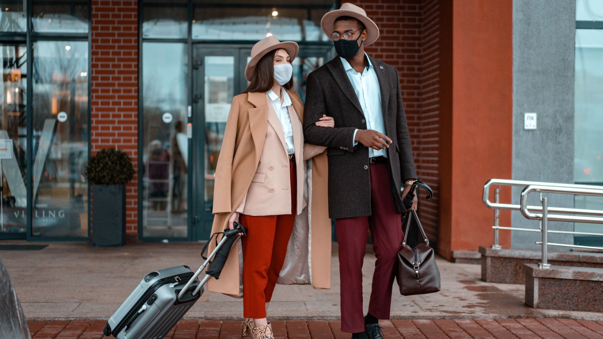 A fashionable couple, wearing face masks, stands with luggage outdoors, ready to travel.