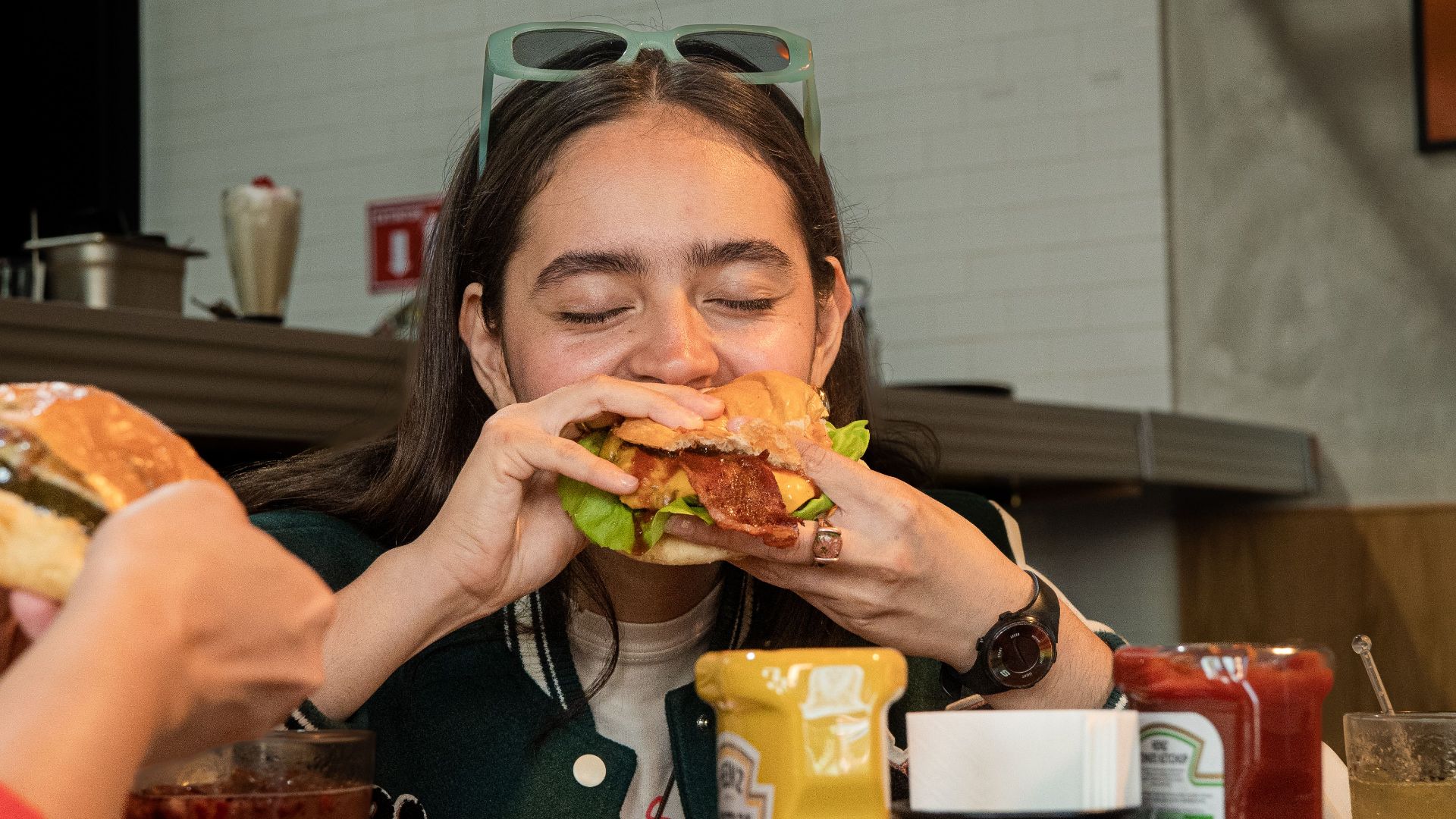 A young woman devours a juicy burger in a casual restaurant setting, surrounded by condiments.