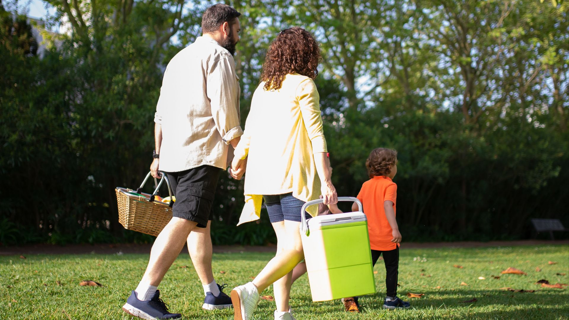 A family with a child carrying picnic essentials walking in a sunny park.