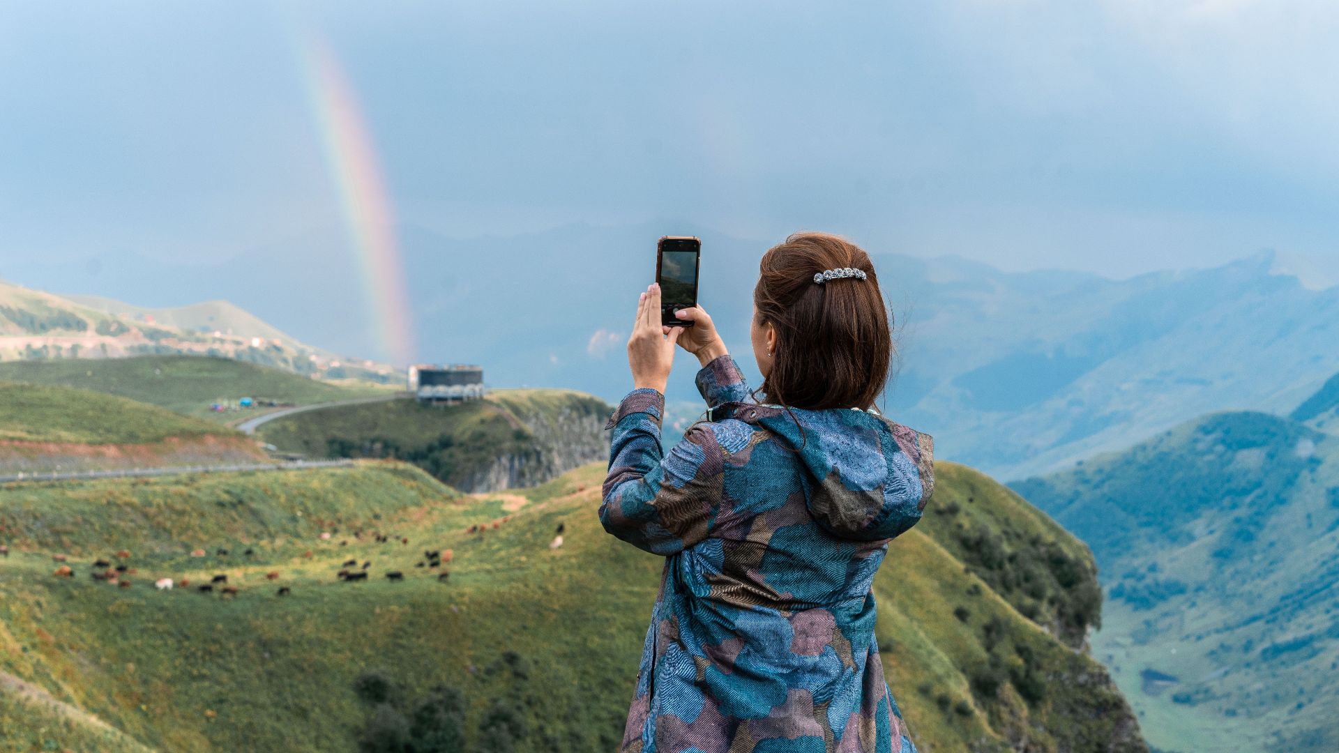 A woman photographs a rainbow over Ganisi, Georgia's scenic mountains.
