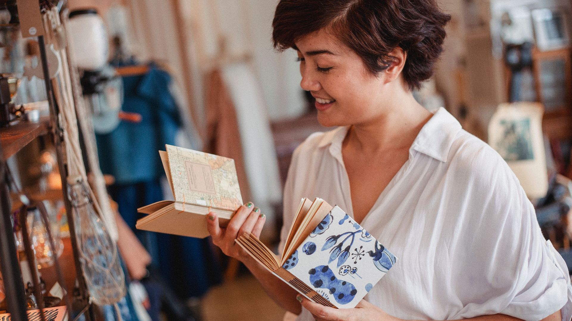 Side view of crop young happy Asian lady choosing various notepads while standing near shelf with stylish goods in vintage boutique