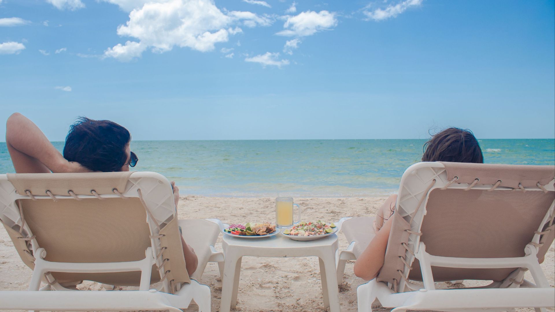 Two people relax on sunbeds with refreshments by the sea, enjoying a sunny summer day.