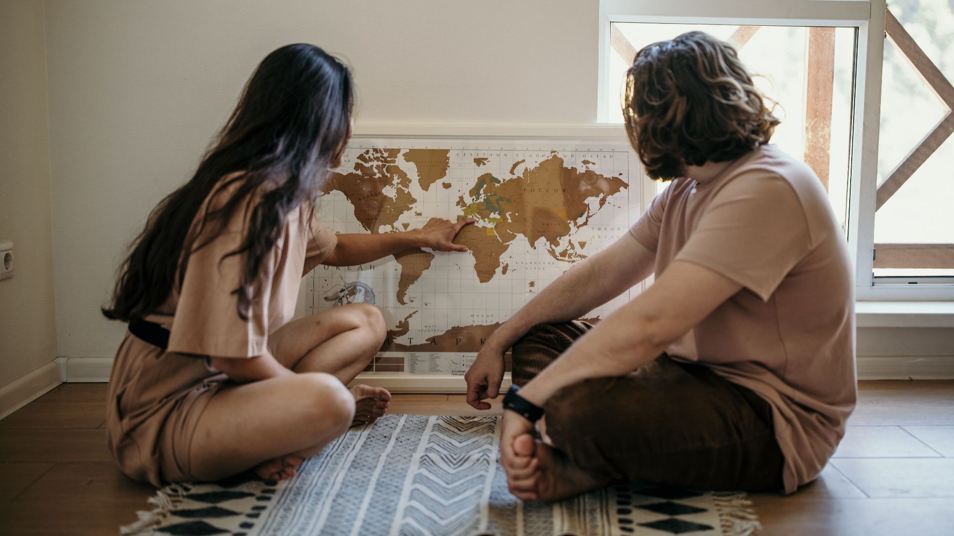 Couple sitting on floor pointing at a world map, planning their next travel adventure indoors.