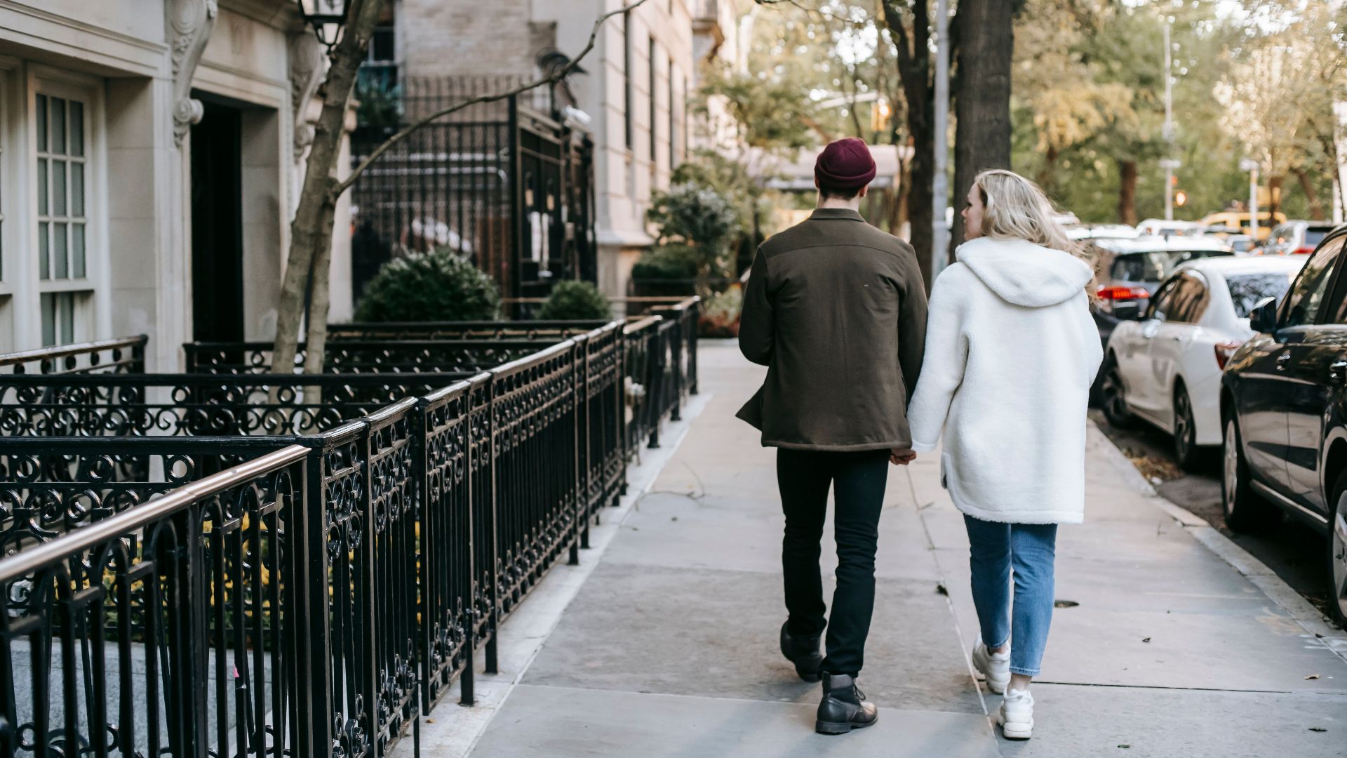 Back view of anonymous trendy young couple in stylish warm clothes holding hands and walking in city street near residential houses in autumn
