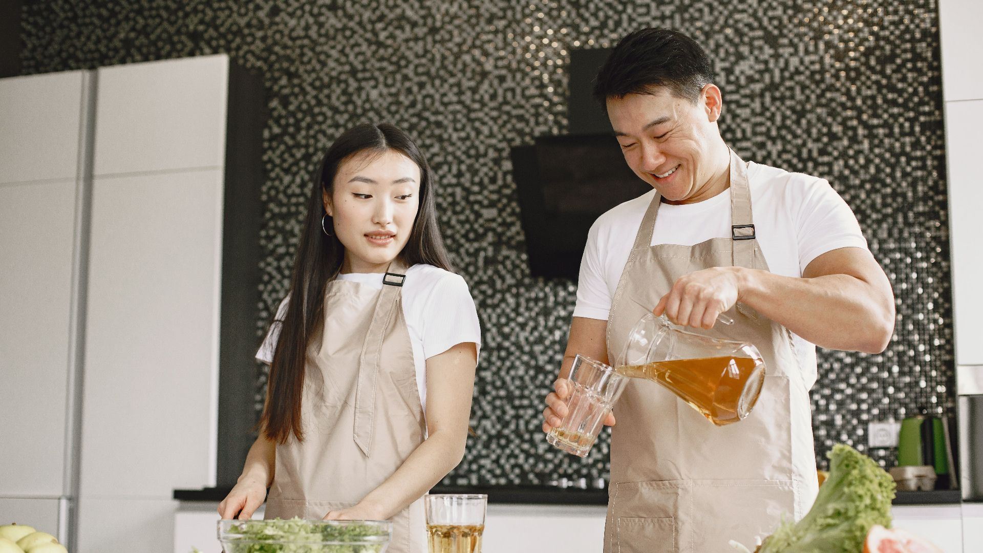 Asian couple preparing salad and drink together in a stylish kitchen, enjoying quality time.