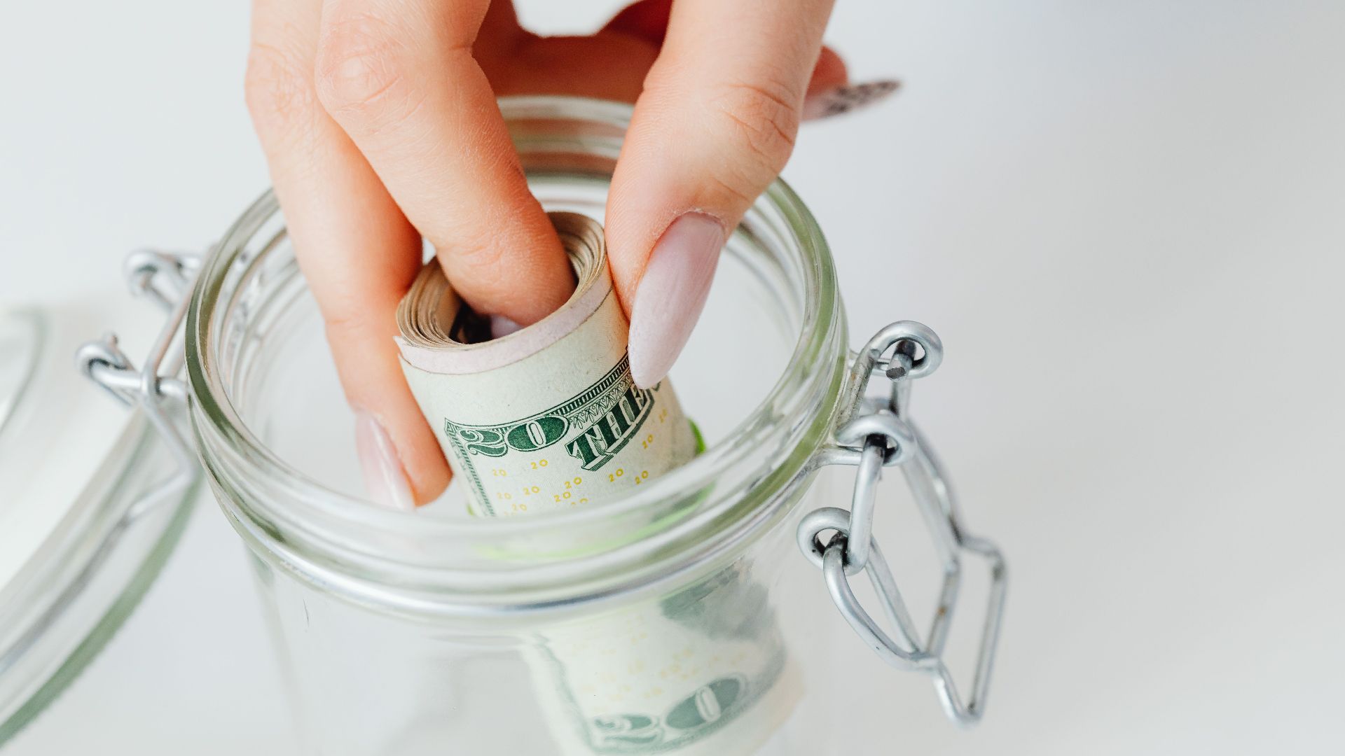 A close-up of a hand placing rolled dollars into a glass jar, symbolizing savings.