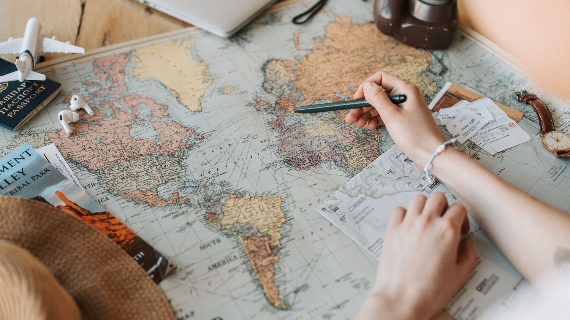Hands marking destinations on a world map surrounded by travel essentials like passport and camera.