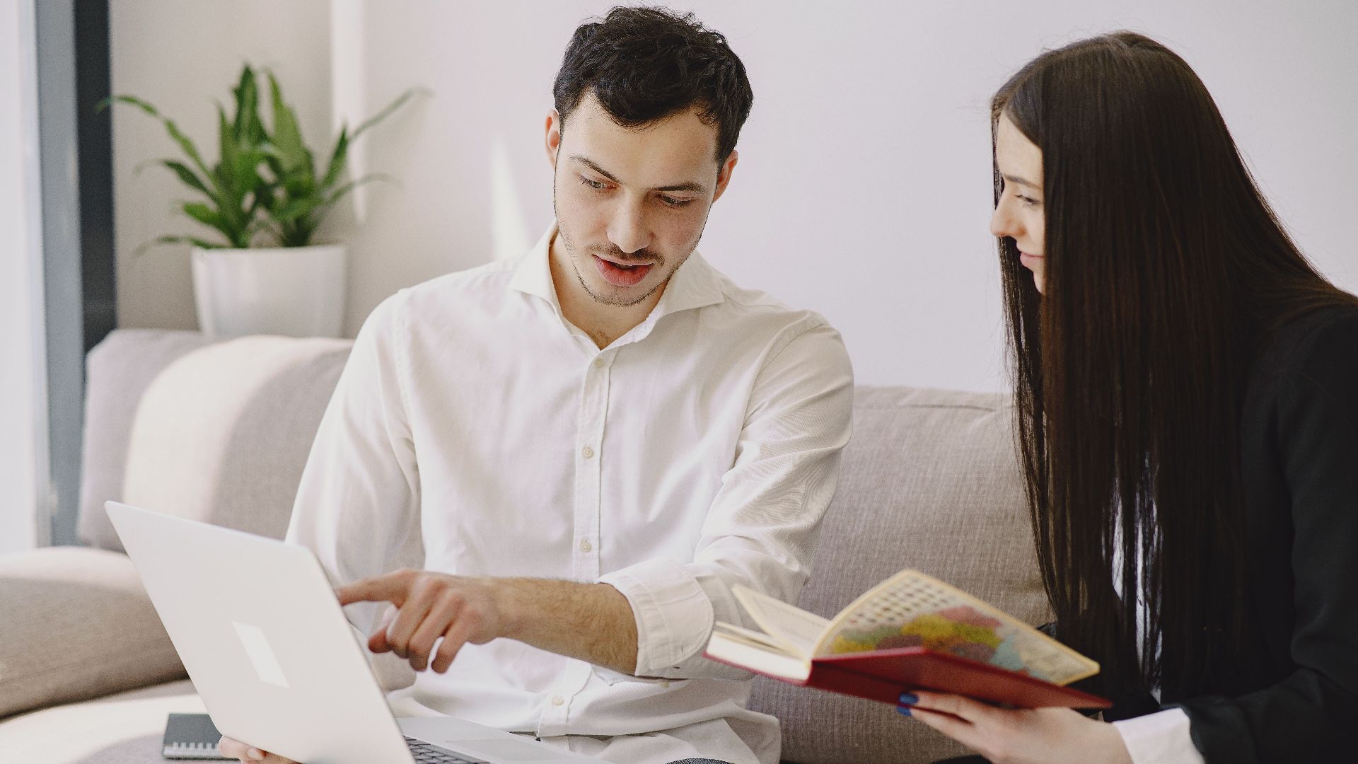 Male worker in formal wear showing with finger at laptop while discussing project with female partner with open copybook and sitting on sofa