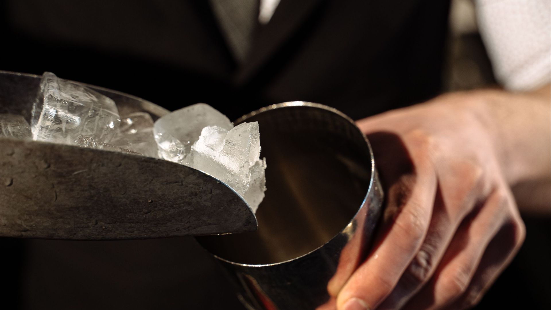 Bartender adds ice into a shaker using a scoop in a dimly lit bar setting.