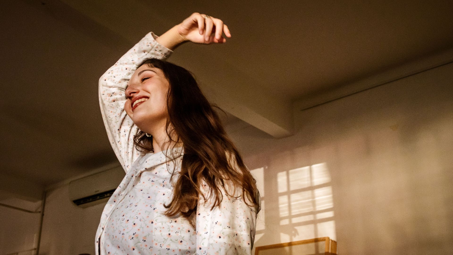 A woman in a light blouse joyfully poses indoors with warm, soft lighting creating a serene mood.
