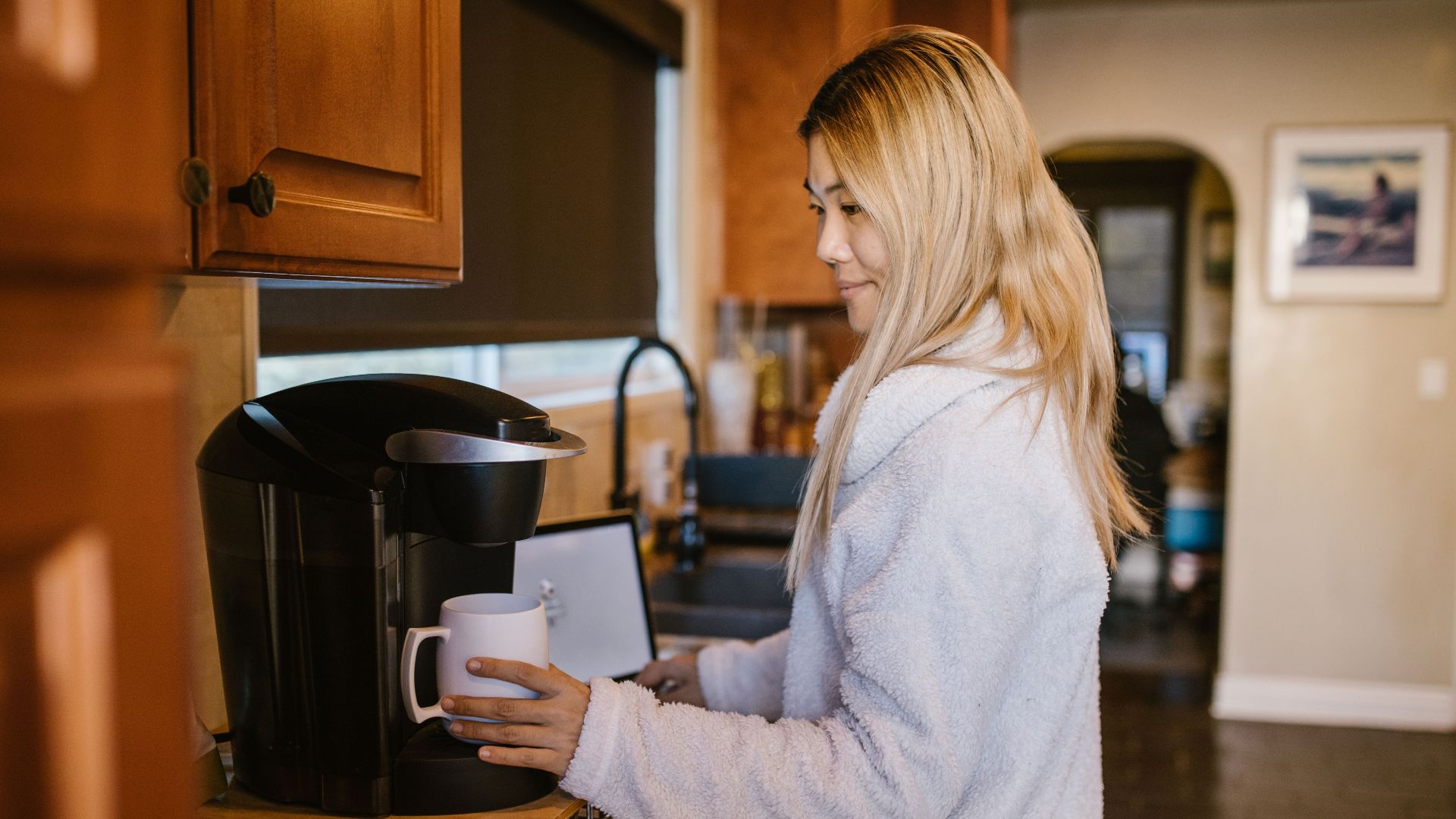 Woman with blonde hair making coffee in a cozy home kitchen setting.
