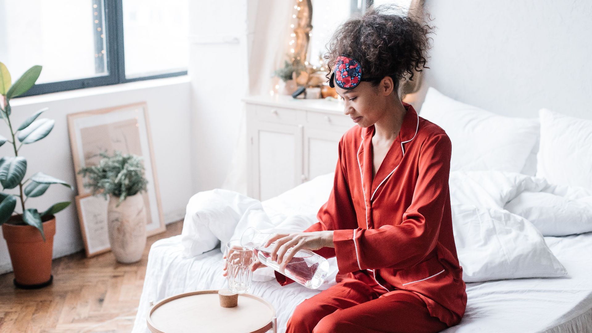 A woman in red pajamas pours water from a carafe into a glass, sitting on a bed in a cozy bedroom.
