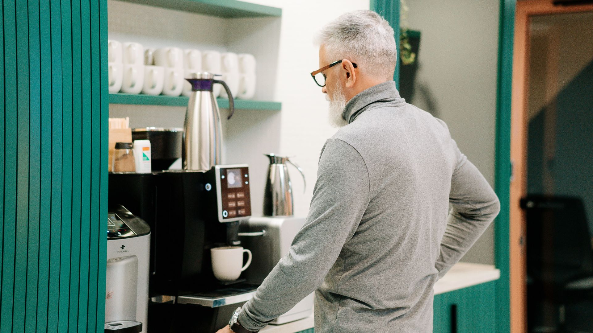 Elderly man in gray sweater making coffee indoors with a modern machine.