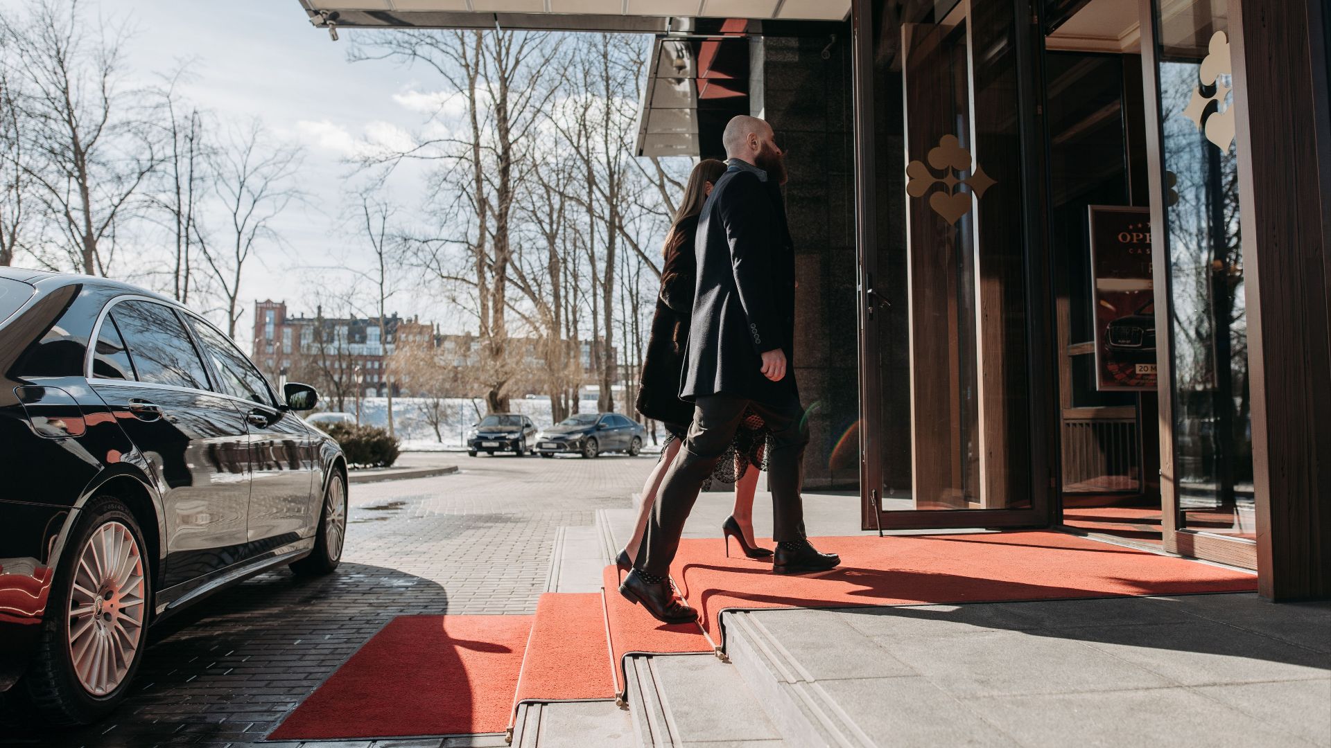 A man and woman elegantly dressed enter a luxury hotel, walking on a red carpet.