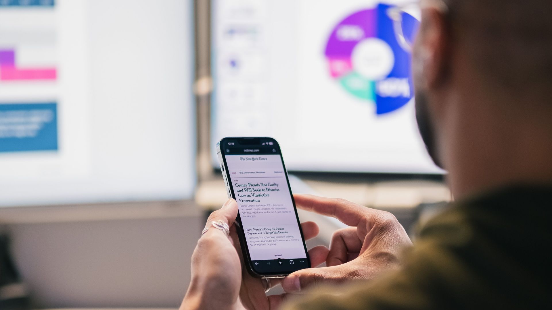 A business professional uses a smartphone while analyzing data on computer screens in a modern office.
